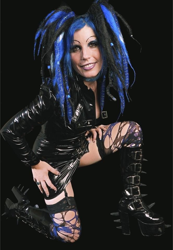 Cybergoth woman with blue dreadlocks, black latex outfit, and spiked boots kneeling.