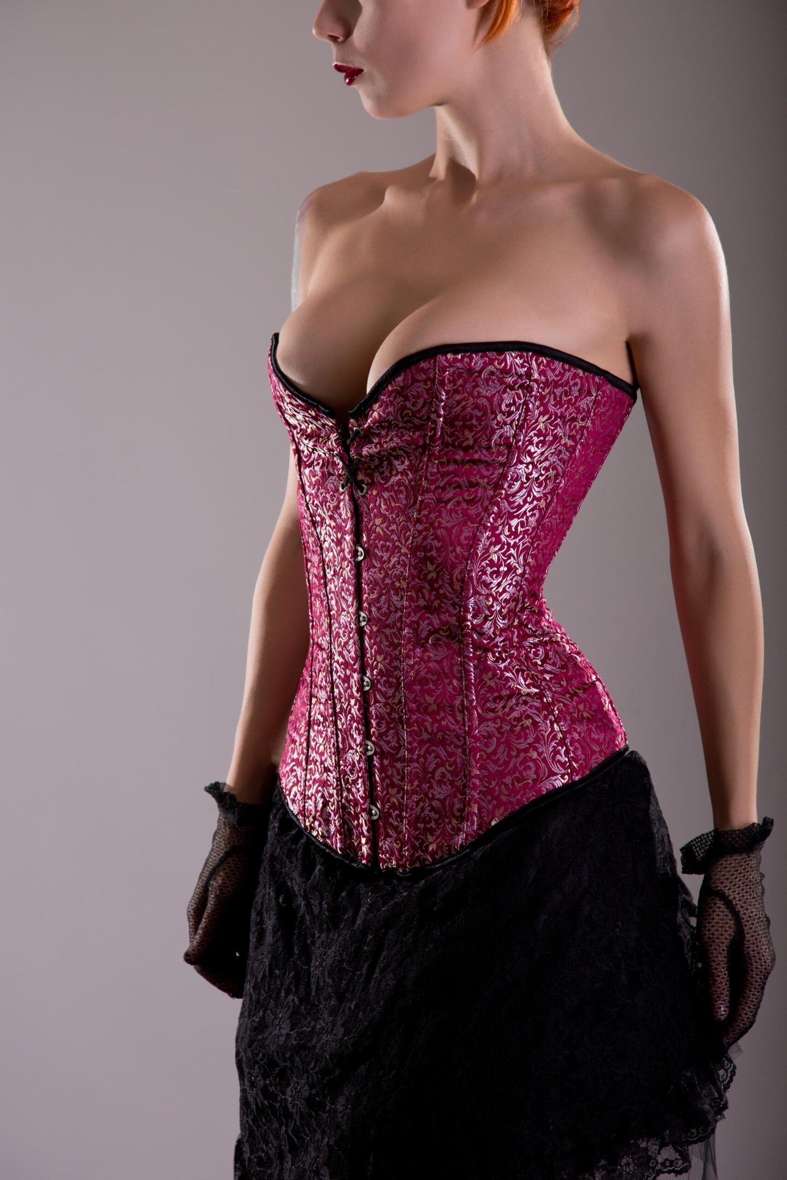 Woman in a pink brocade overbust corset with black lace posing in profile.