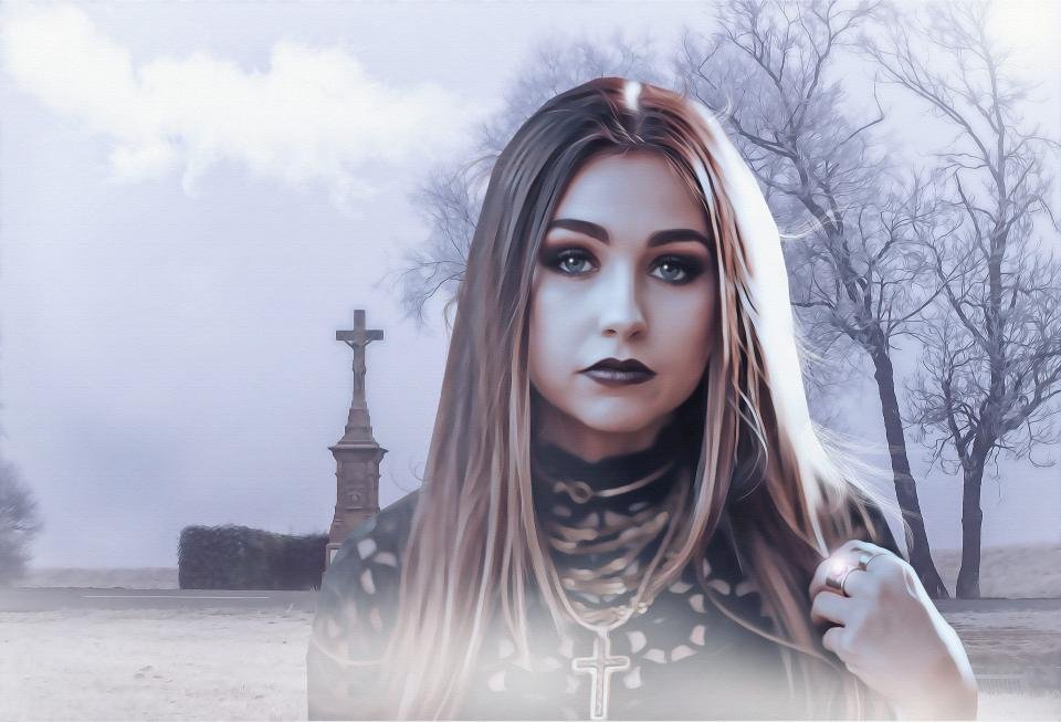 Gothic portrait of a woman wearing a black lace top and cross necklace, standing outdoors near a graveyard in a pale, misty setting.