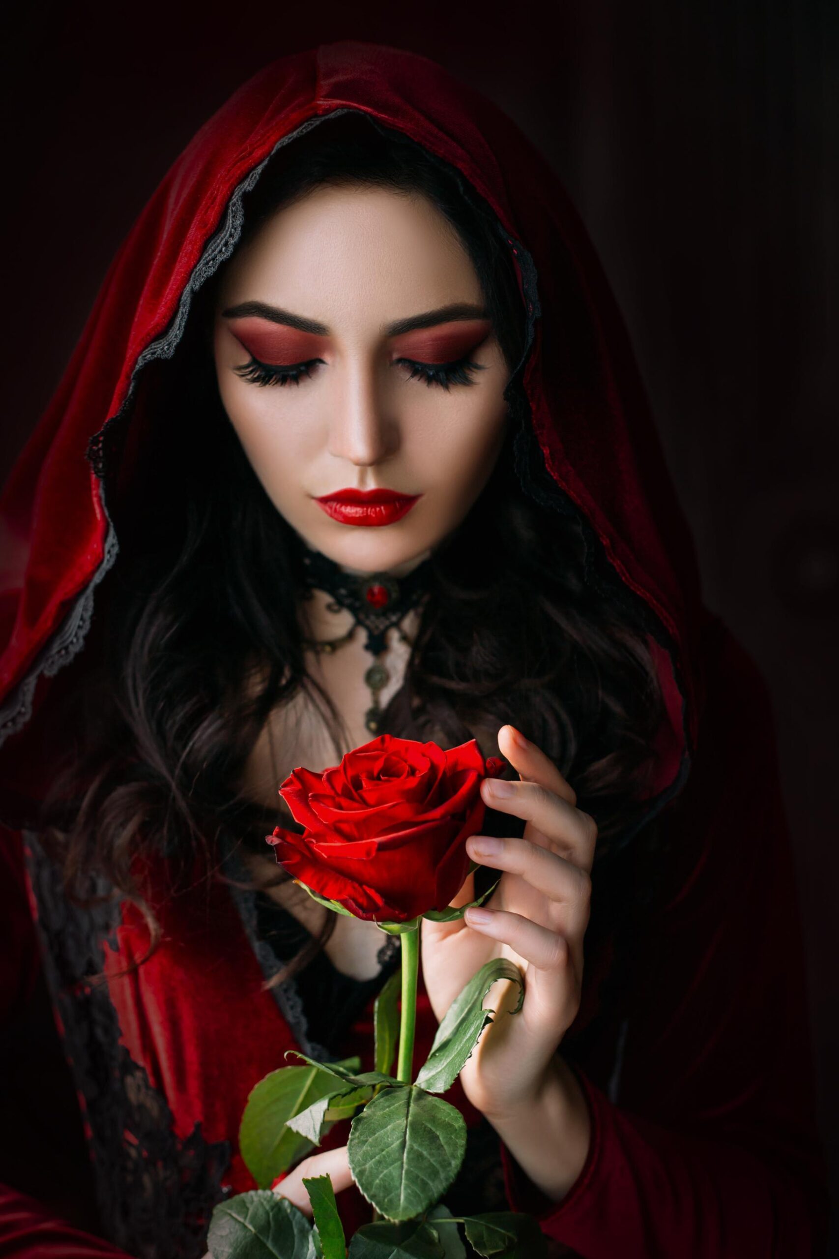 Gothic woman in red hood holding a red rose, eyes downcast, dark romantic styling, black background, dramatic lighting.