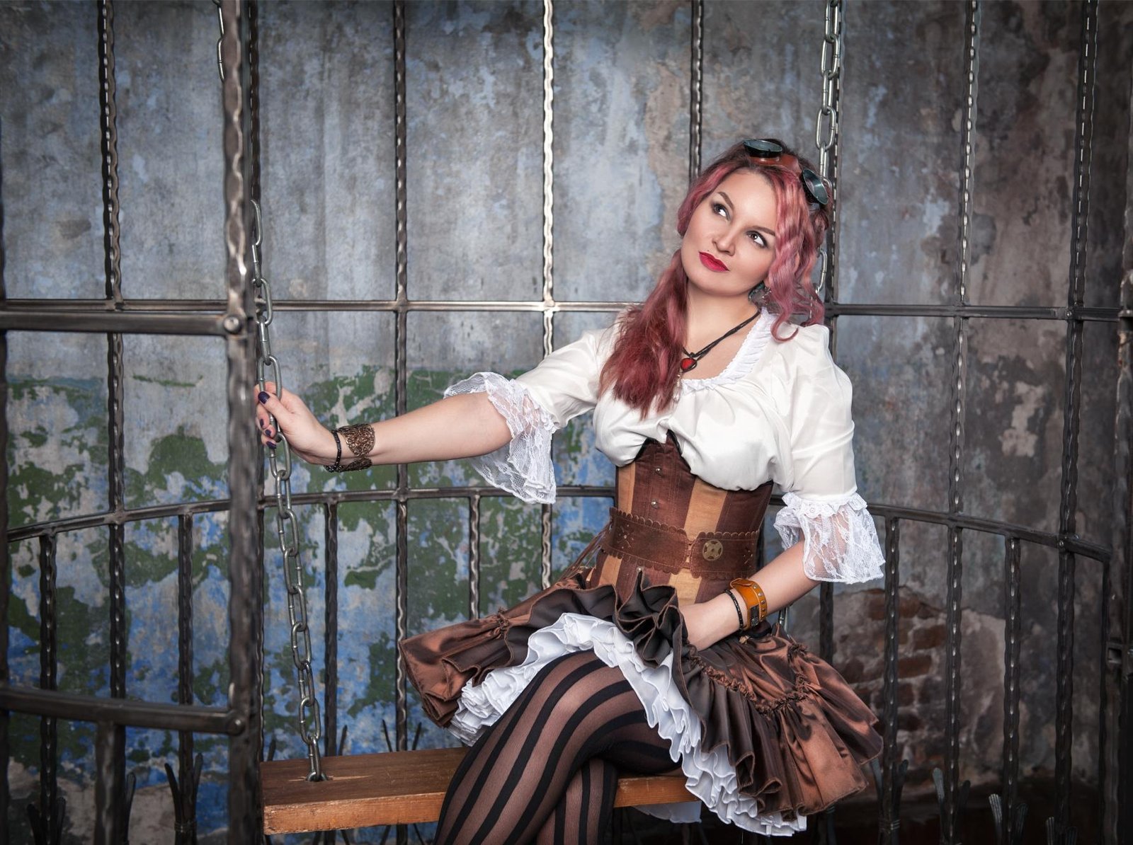 Woman in steampunk-inspired outfit sitting on a swing inside a metal cage, wearing corset dress and striped stockings.