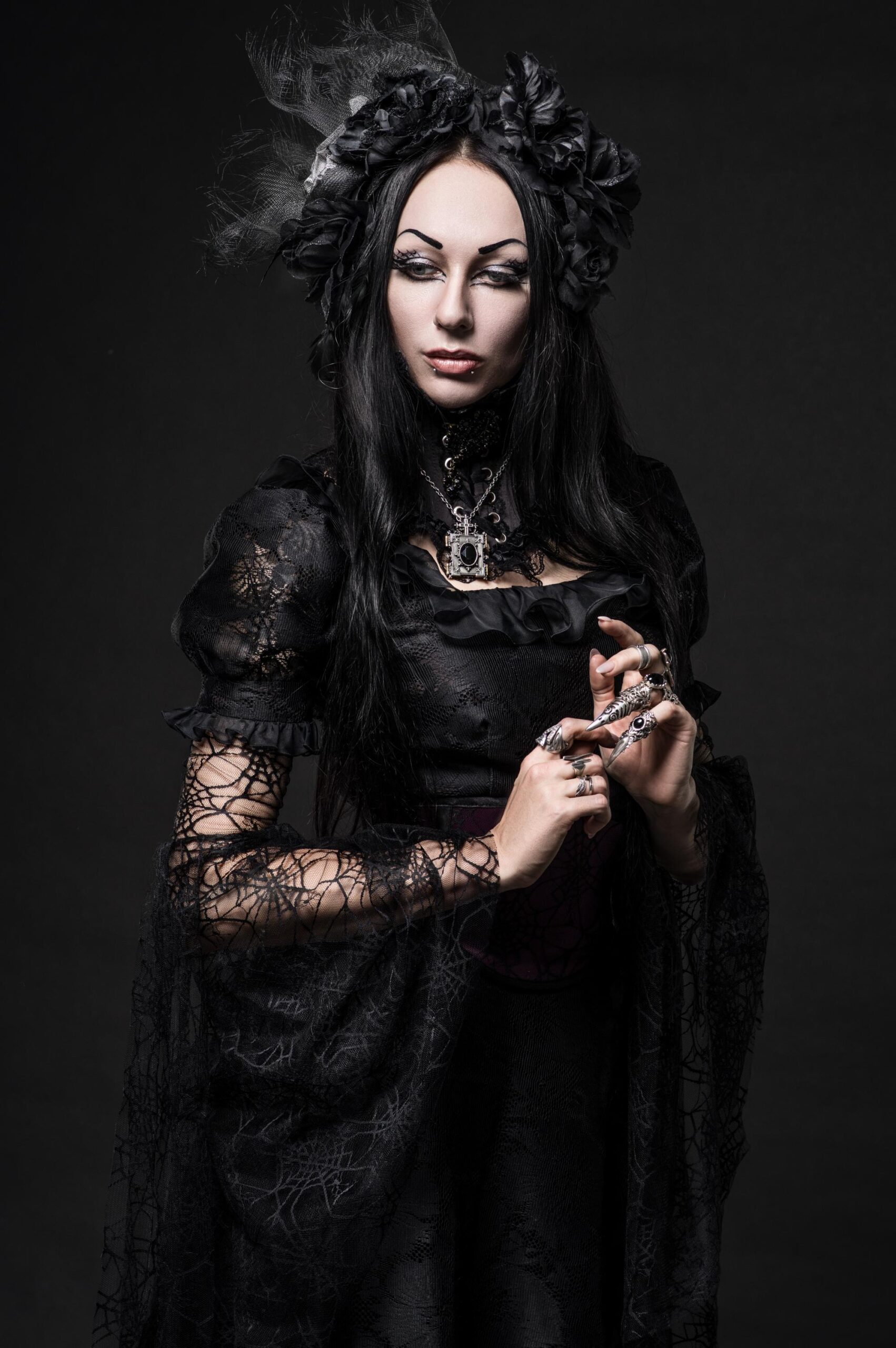 Victorian gothic woman in black lace dress with ornate jewelry and headpiece, pale skin, dark makeup, posed against a dark studio background.