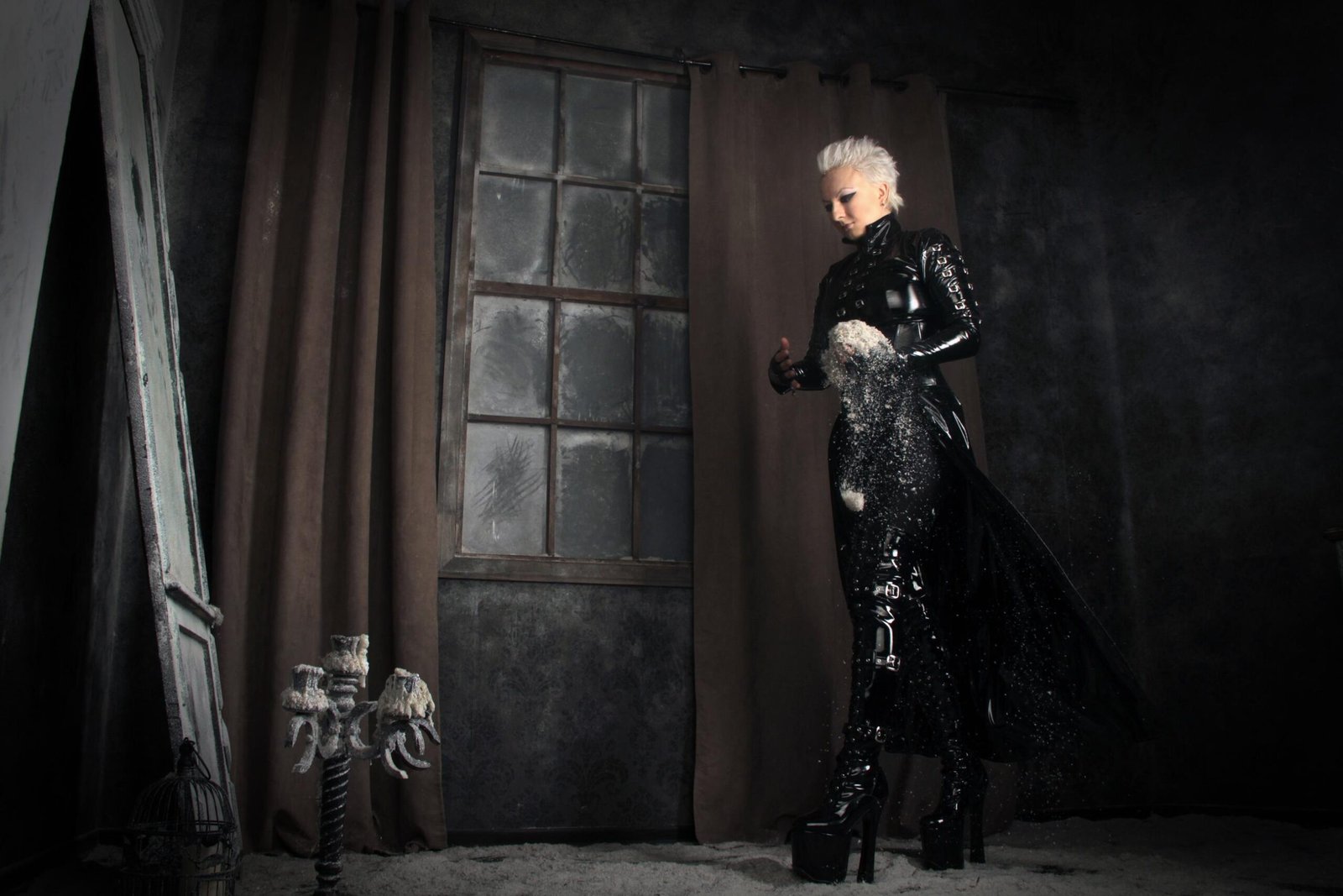 White-haired woman in a long black latex coat standing in a dark room with curtains and window, holding crumbling white dust.