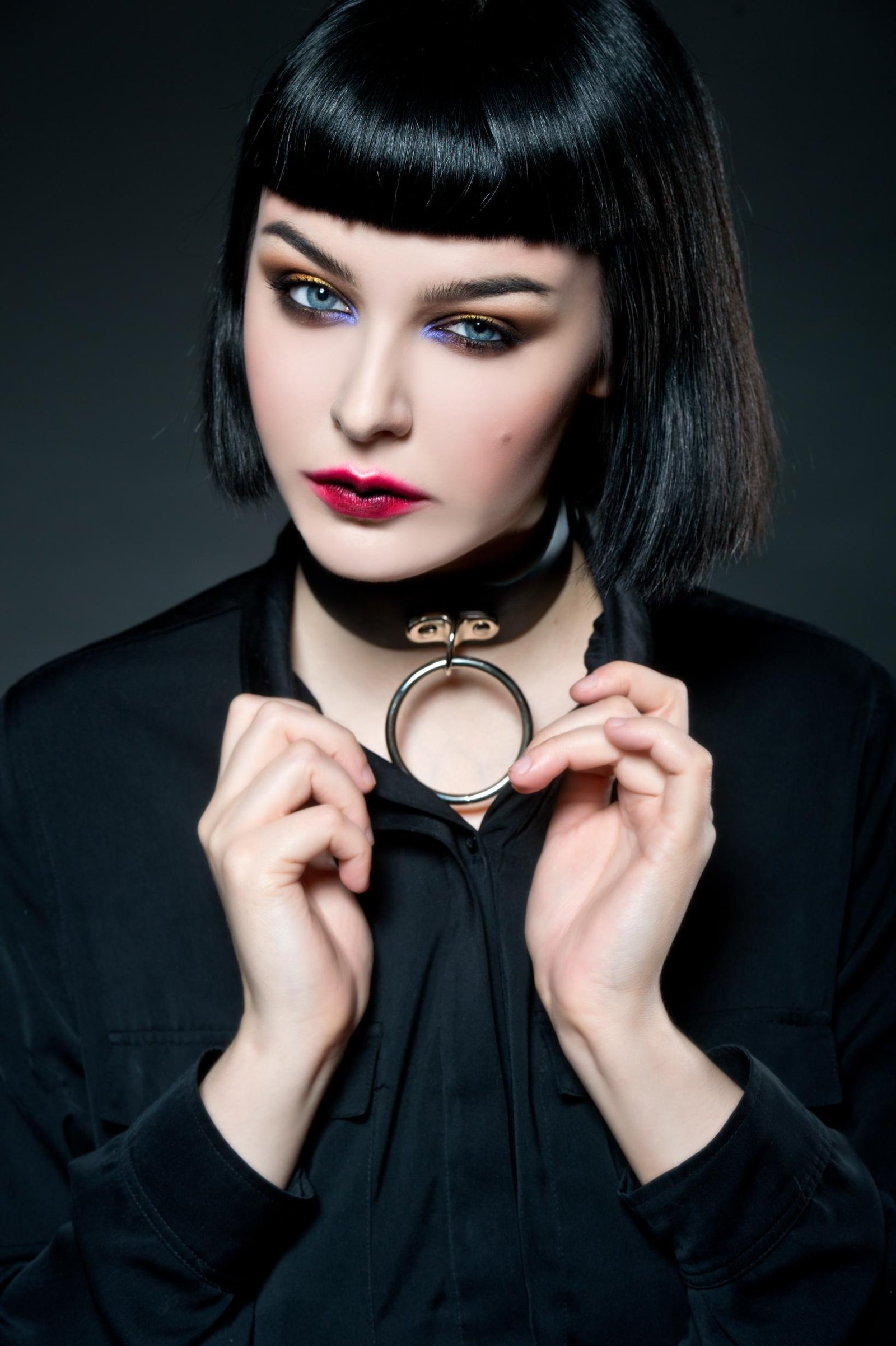 Studio portrait of a pale gothic woman with short black bob, blunt bangs, red lipstick, and a black choker with metal ring.
