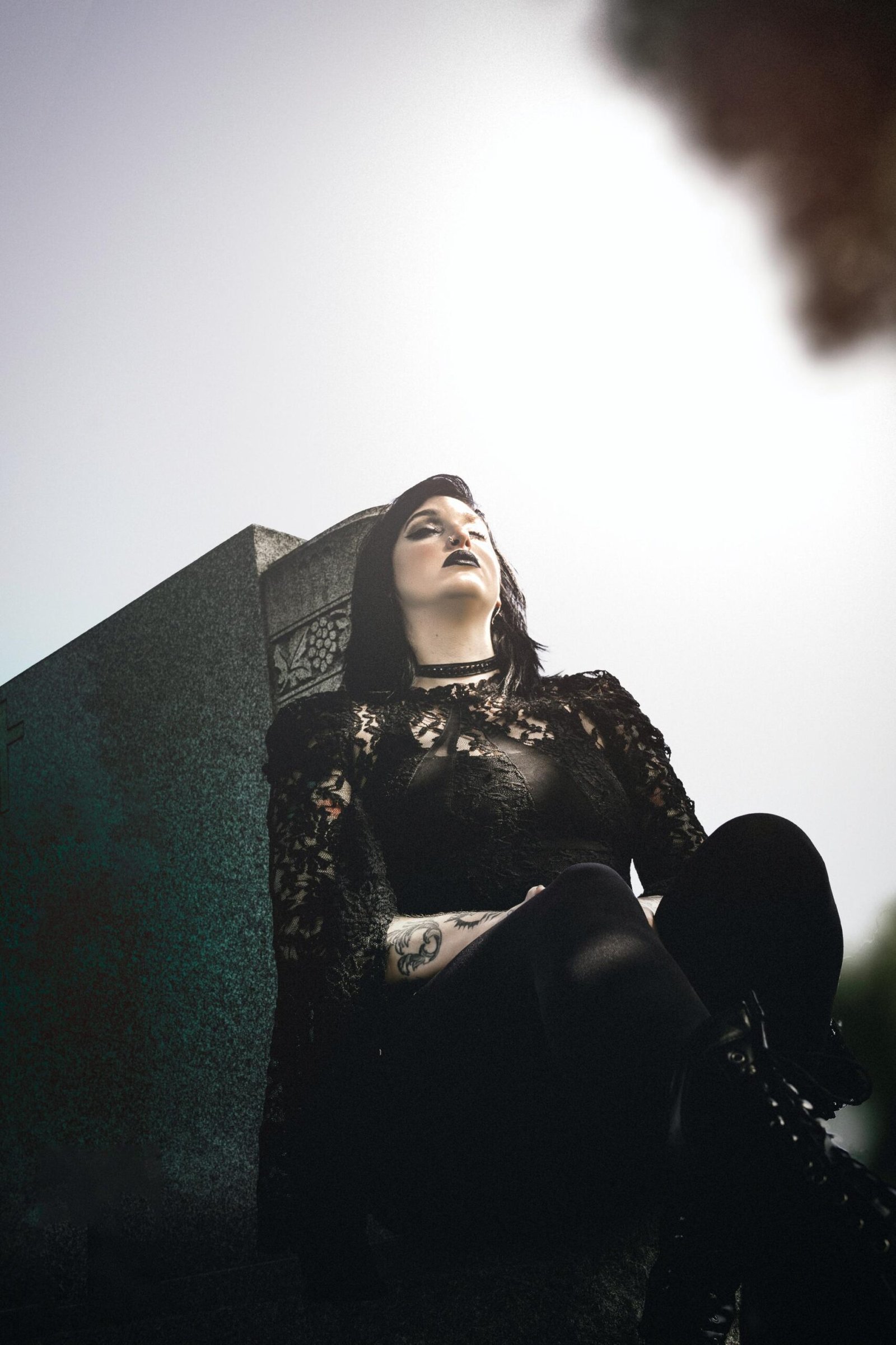 Gothic woman dressed in black lace sitting on a stone structure outdoors, dramatic low-angle portrait with moody lighting.