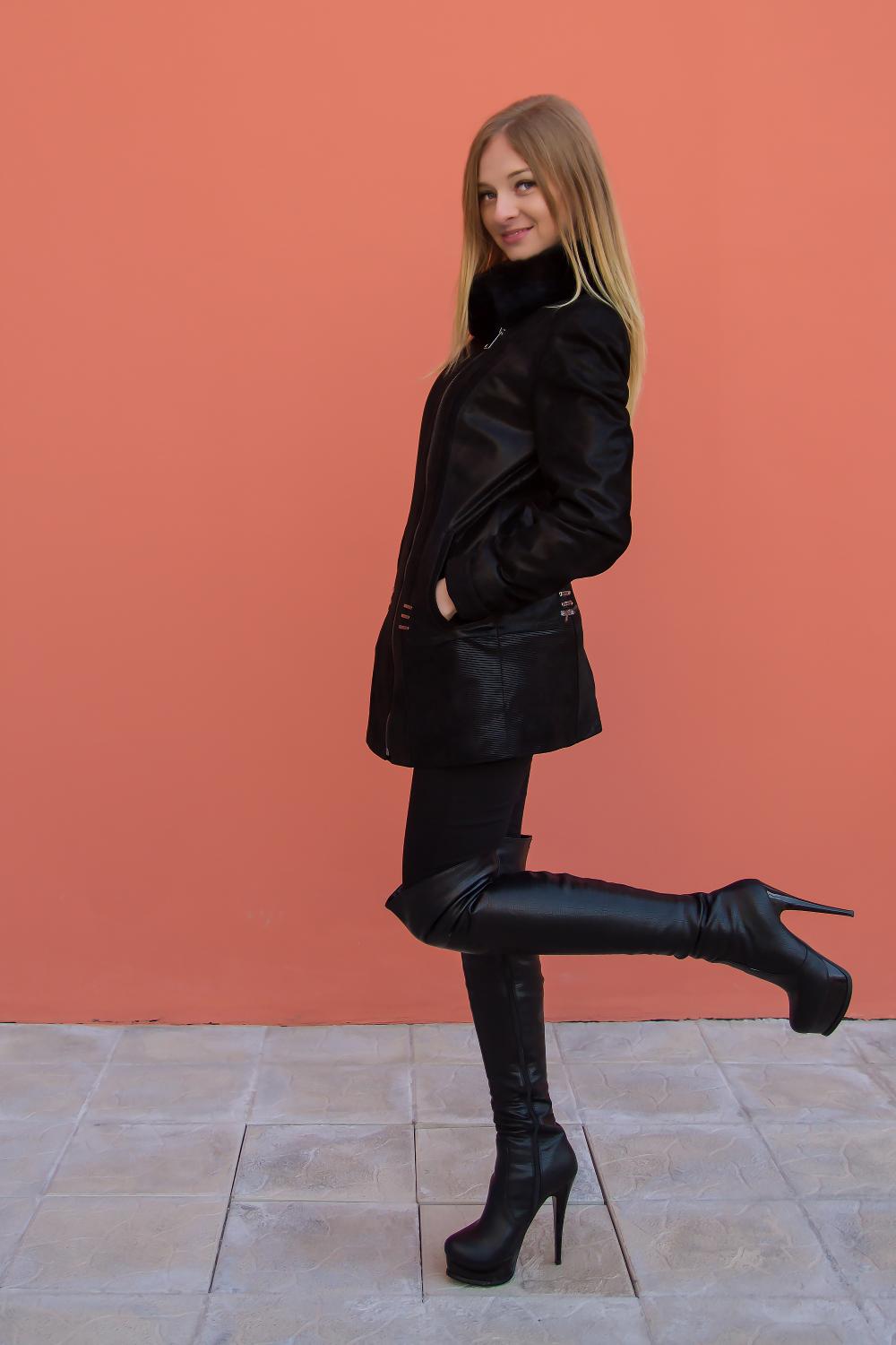 Blonde woman in black leather jacket, leggings, and high heels posing with one leg raised against a coral wall