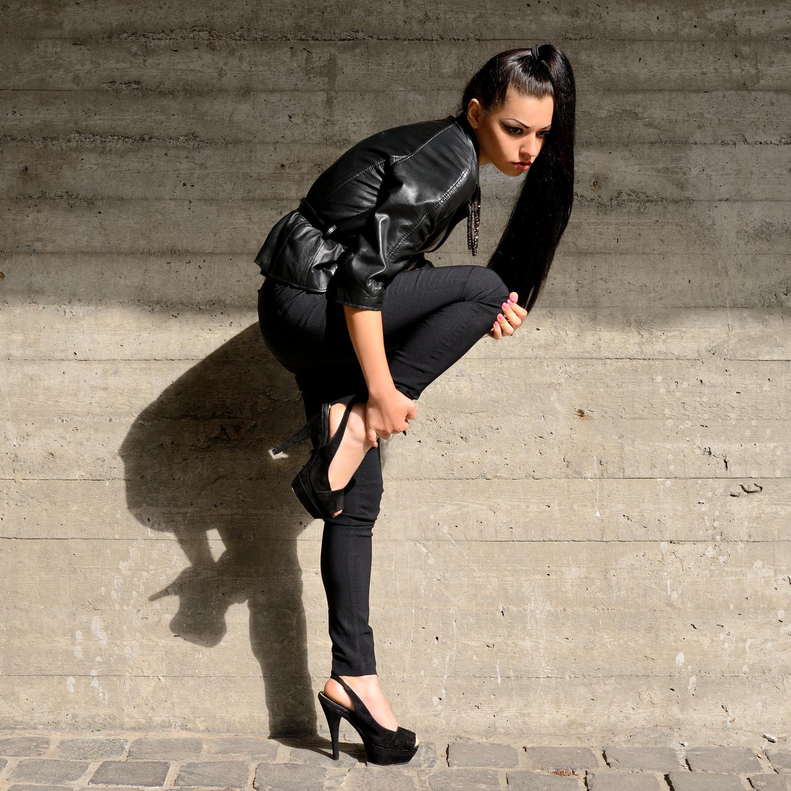 Woman in black leather jacket adjusting high heel against concrete wall, edgy urban fashion portrait