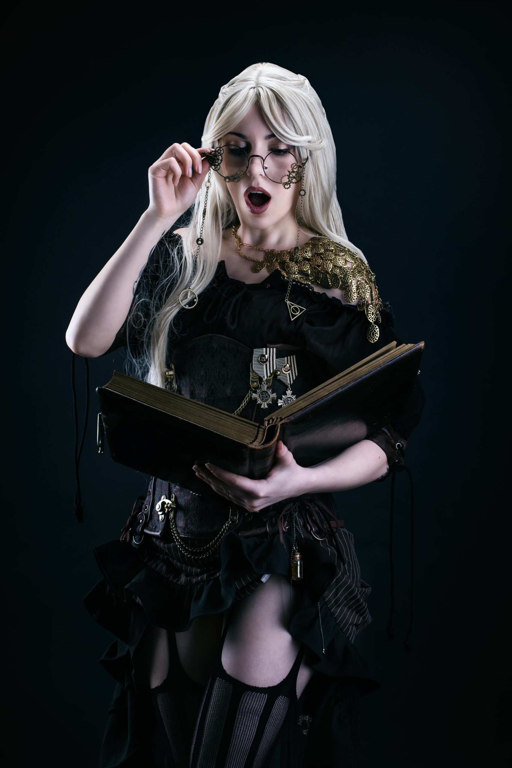 Blonde steampunk woman reading large book, wearing ornate glasses and black Victorian-inspired outfit.