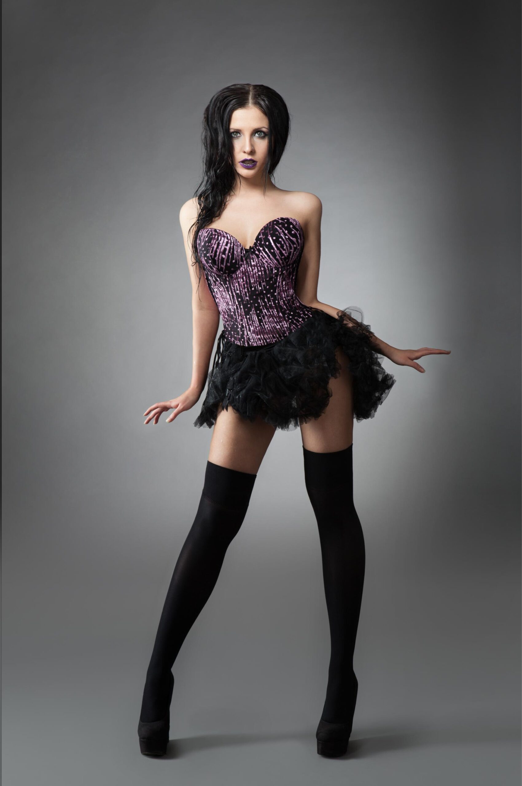 Woman in purple corset, short black skirt and thigh-high stockings posing against gray studio background.