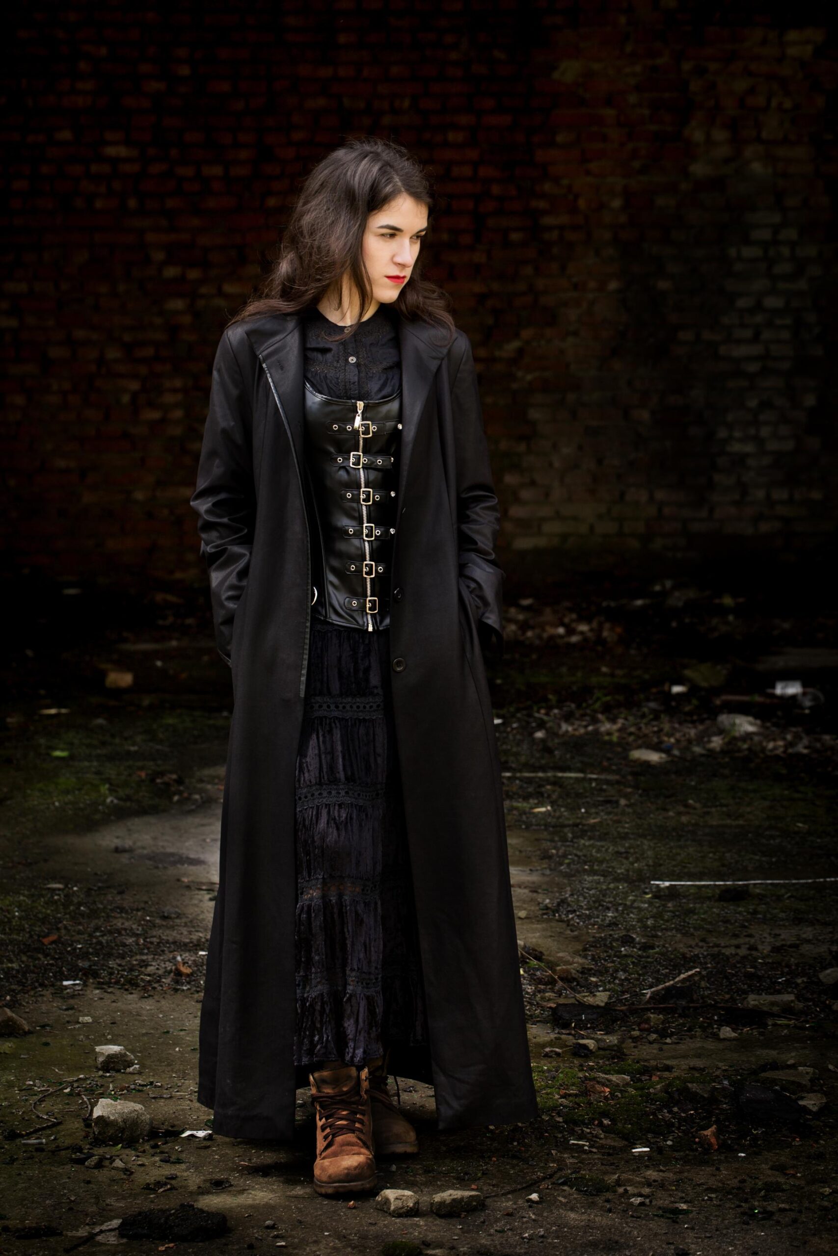 Woman in long black coat and buckled corset standing in abandoned industrial setting.