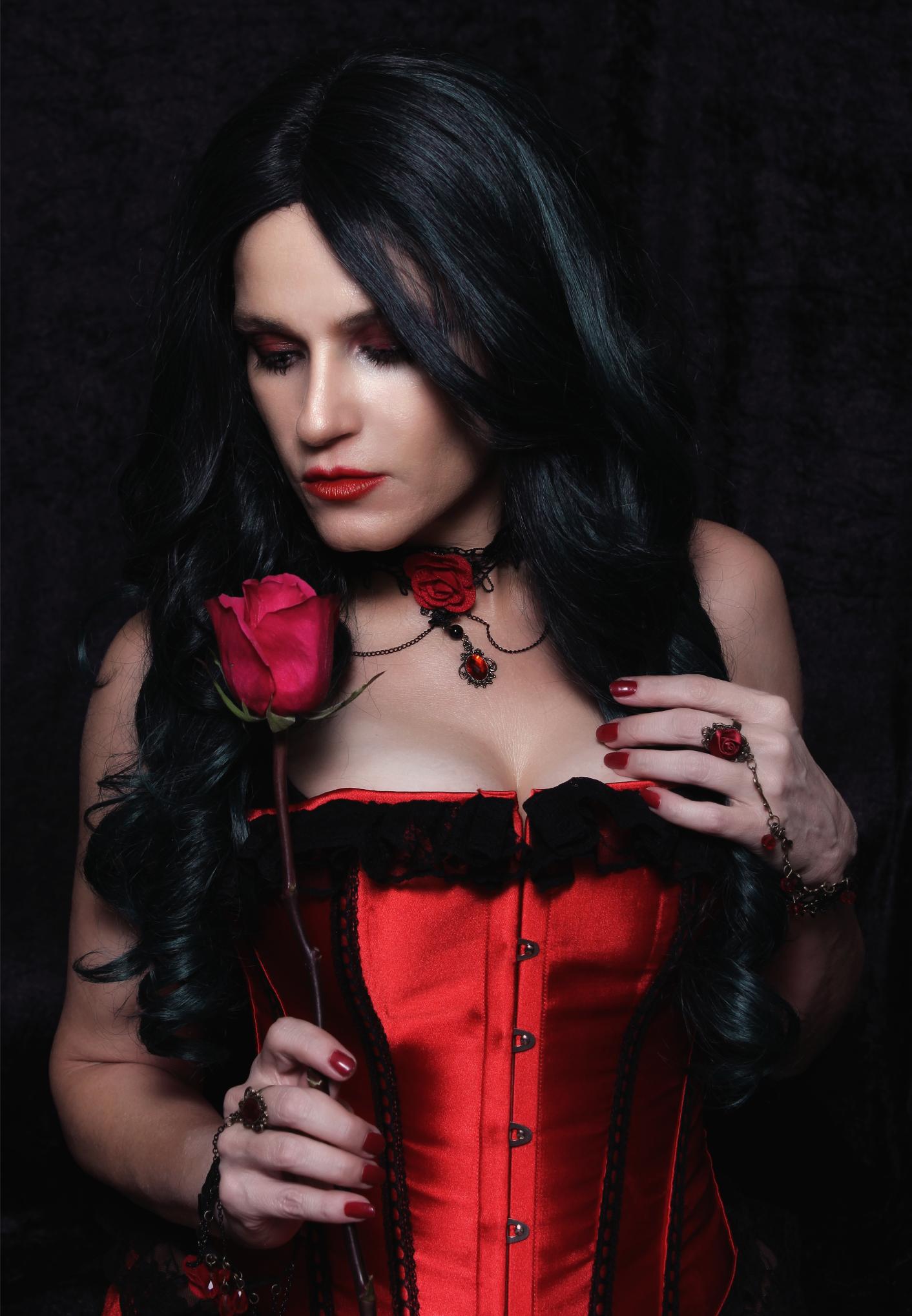 Woman in red corset holding a red rose against dark backdrop, gothic makeup and black hair.