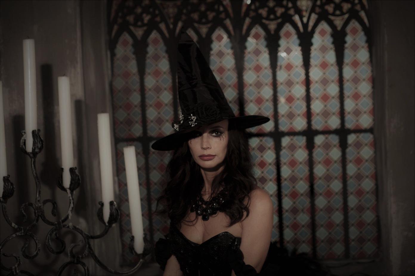 Portrait of woman in black witch hat and dress beside tall candles and stained glass window.