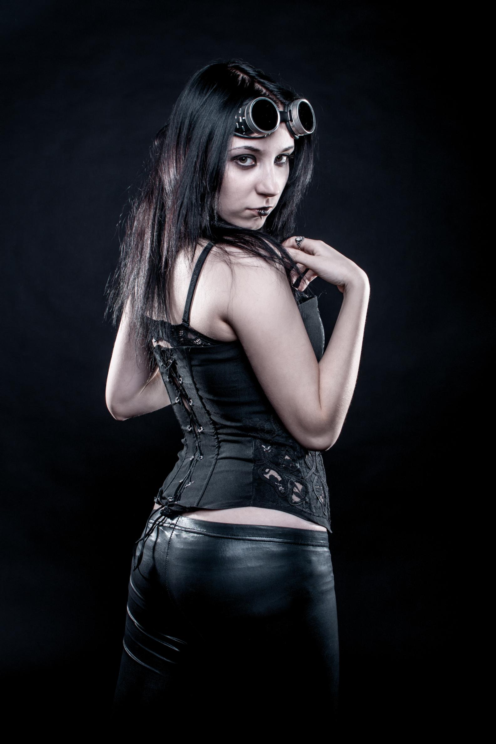 Woman in black corset and leather pants with steampunk goggles, posing over shoulder on dark background.