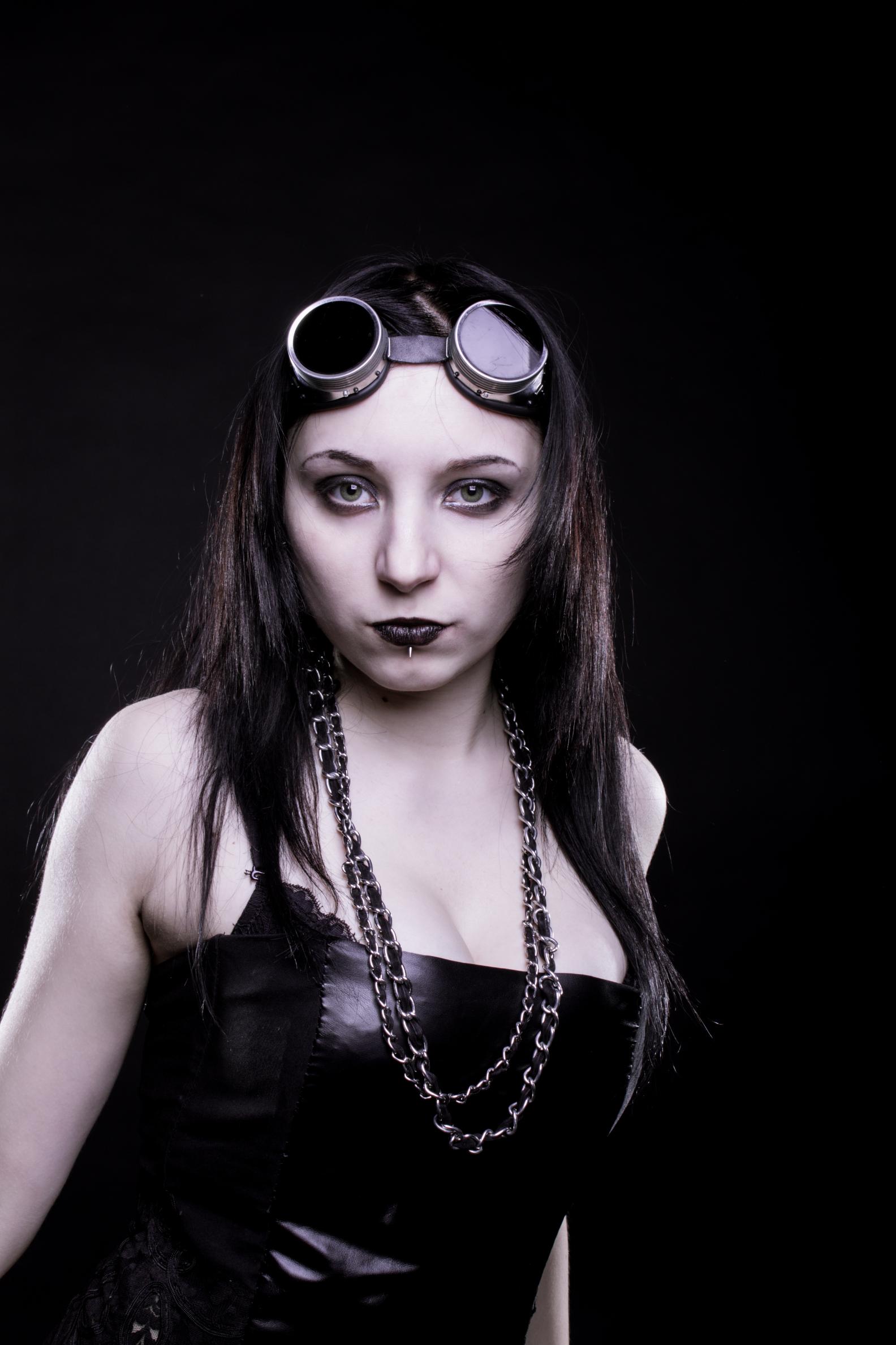 Gothic woman with goggles and chain necklace, black lipstick and corset, staring at camera on dark background.
