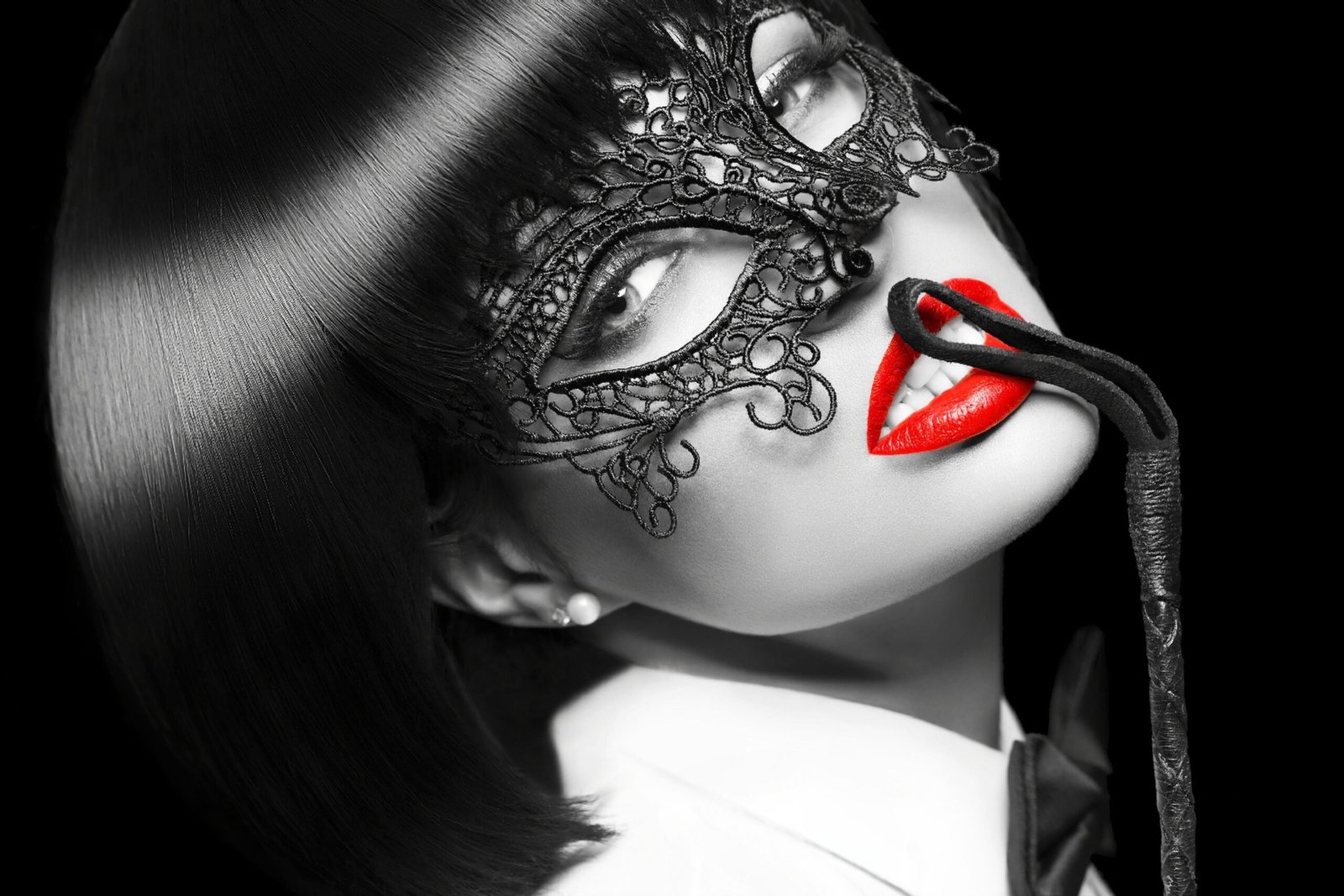 Close-up portrait of woman in lace mask with bright red lips holding a riding crop against a dark background
