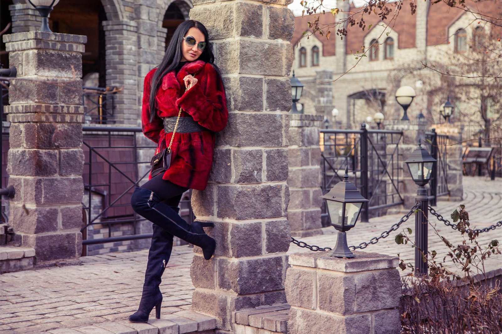 Woman in red fur coat and thigh-high boots leaning against stone pillar in historic city street, stylish winter fashion pose