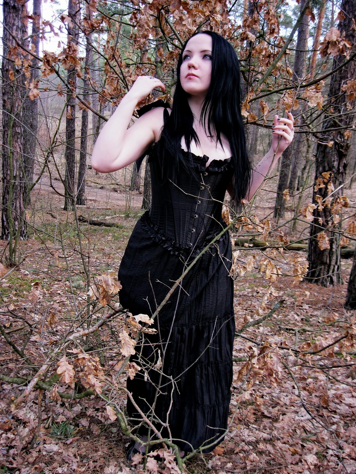 Woman in black corset dress posing among dry leaves and branches in a woodland setting.