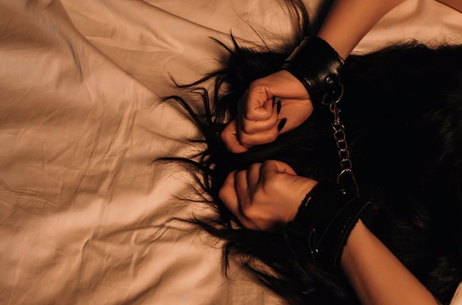 Person lying on a bed with wrists restrained in leather cuffs, hands positioned above their head