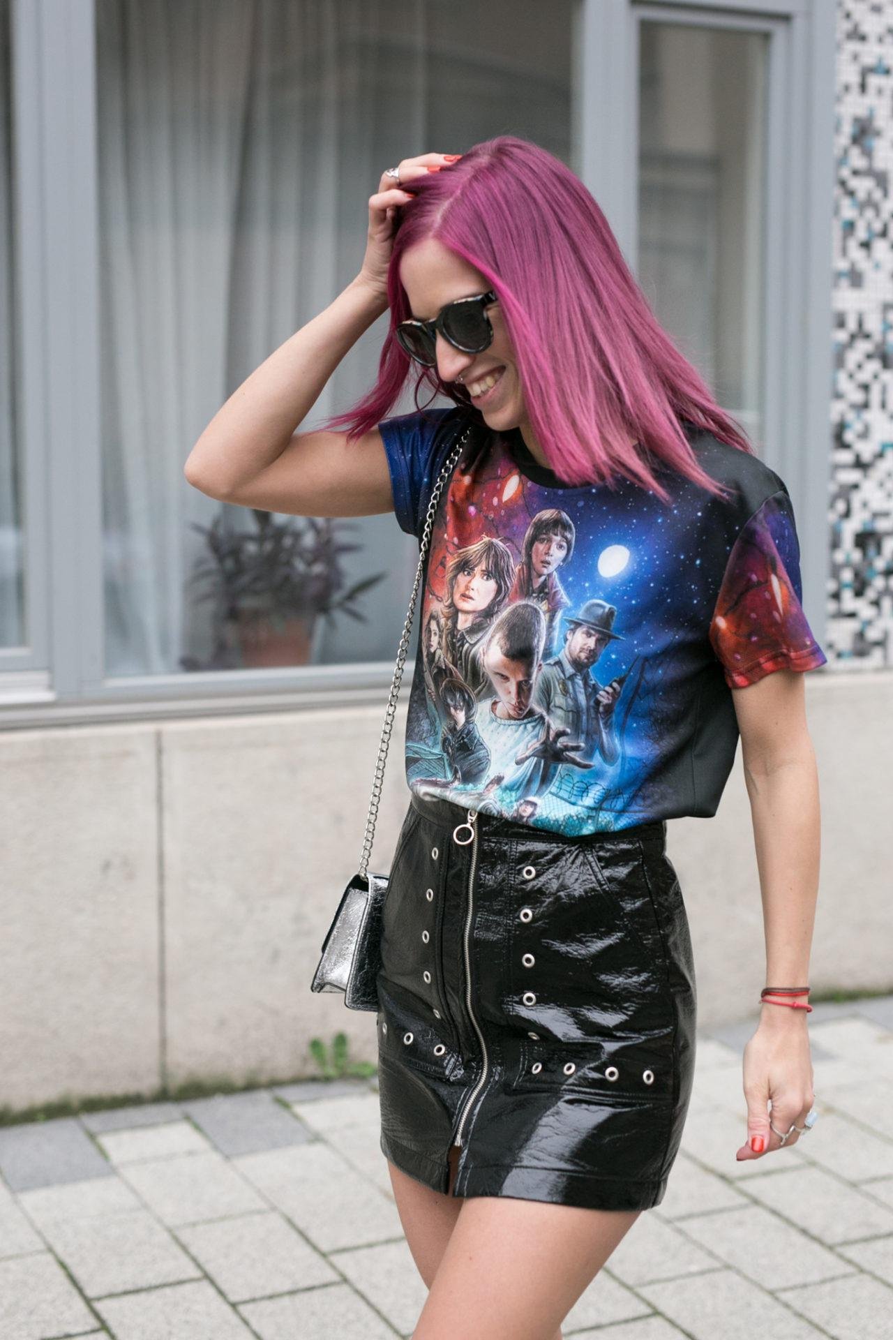 Woman with pink hair wearing graphic tee and black vinyl skirt walking outdoors, casual street style