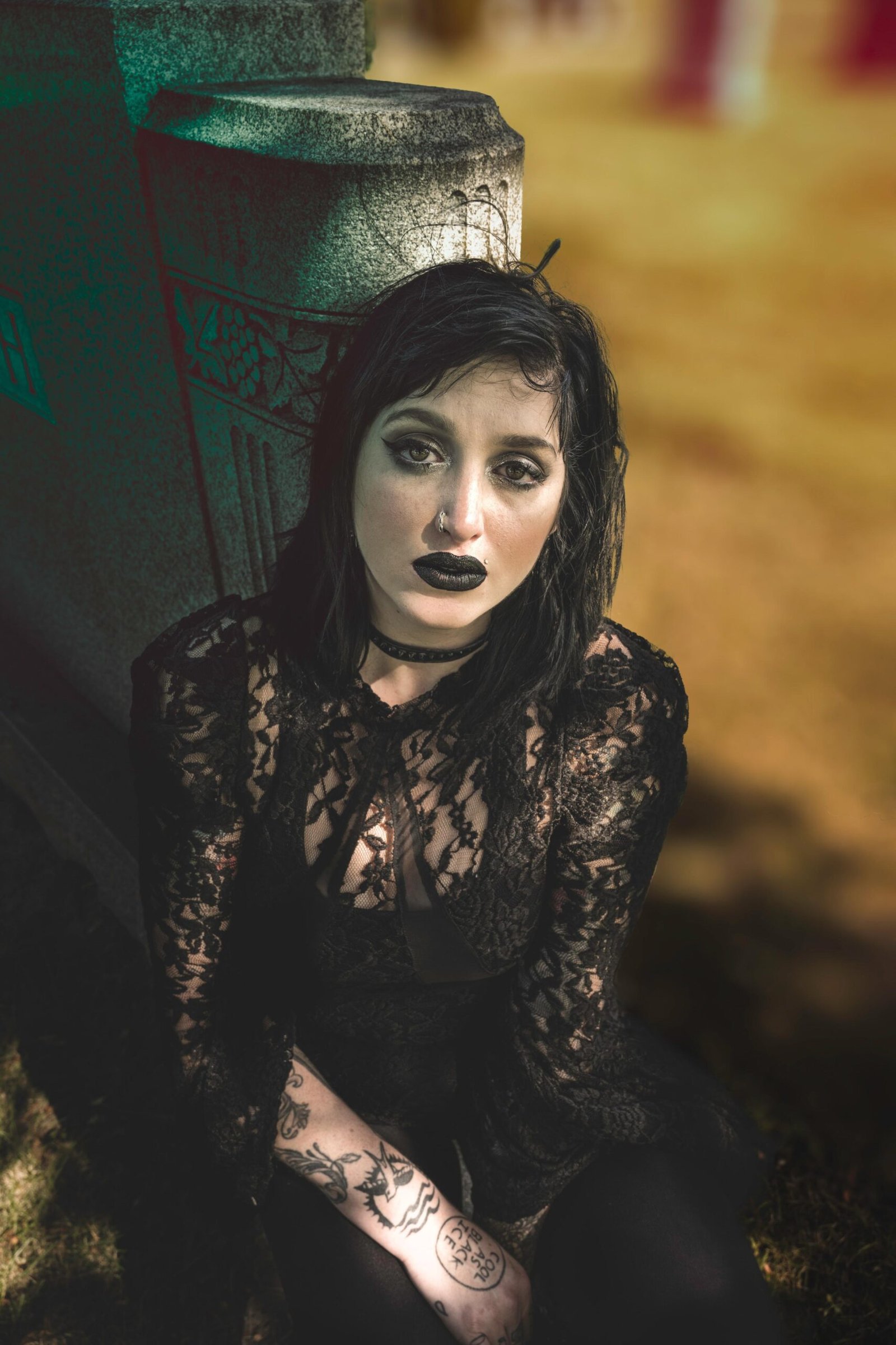 Gothic woman in black lace top sitting by a stone monument outdoors, dark makeup and intense gaze toward the camera.