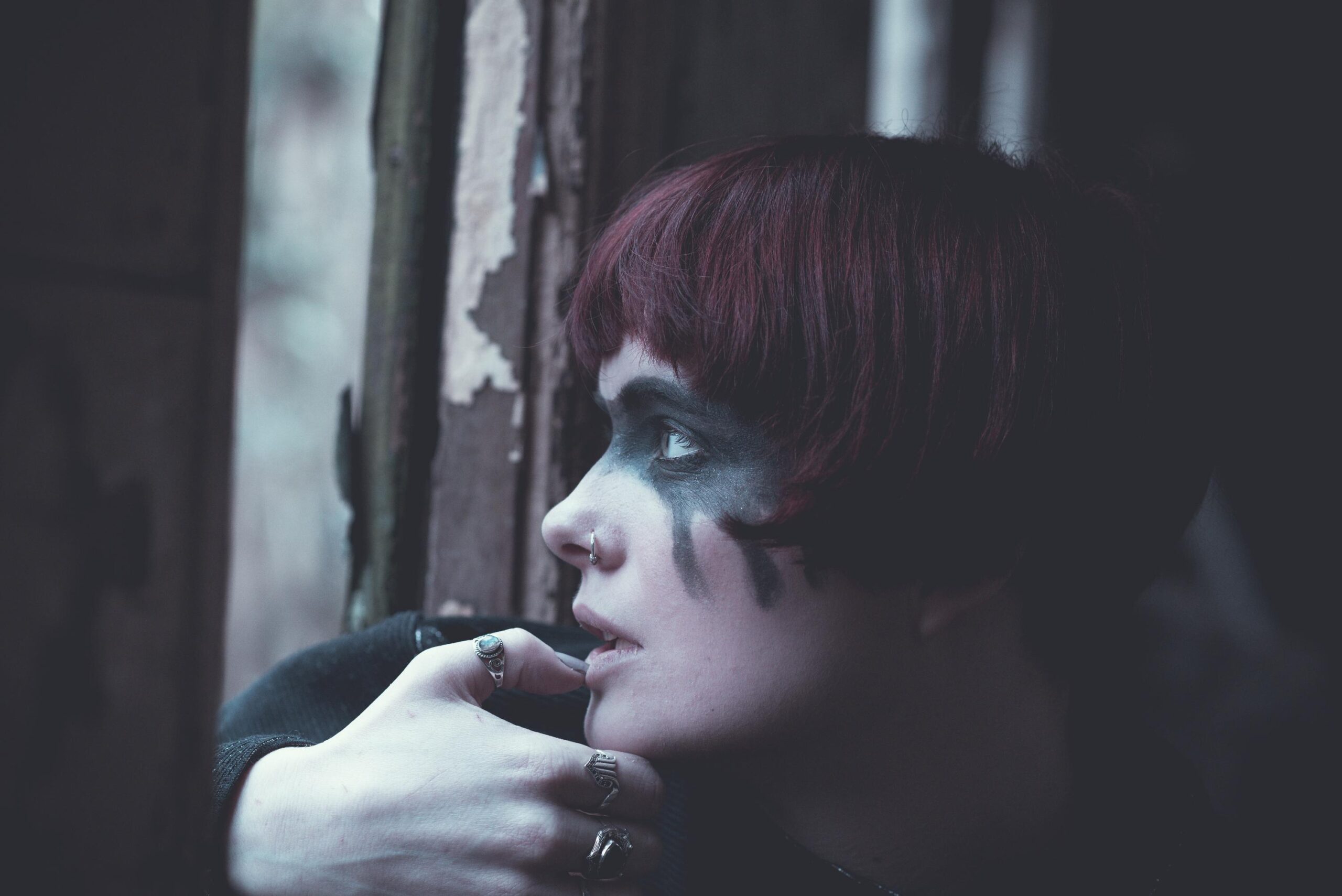 Woman with short red hair and dark eye makeup looking out worn window in moody light.