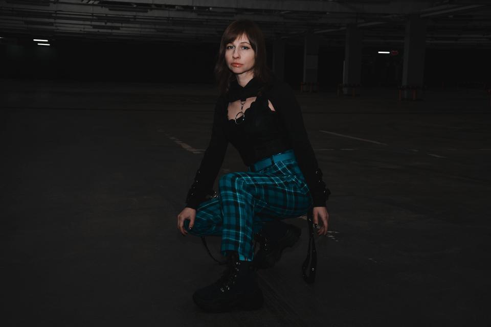 Woman crouching in dark parking garage wearing black top and green plaid pants.