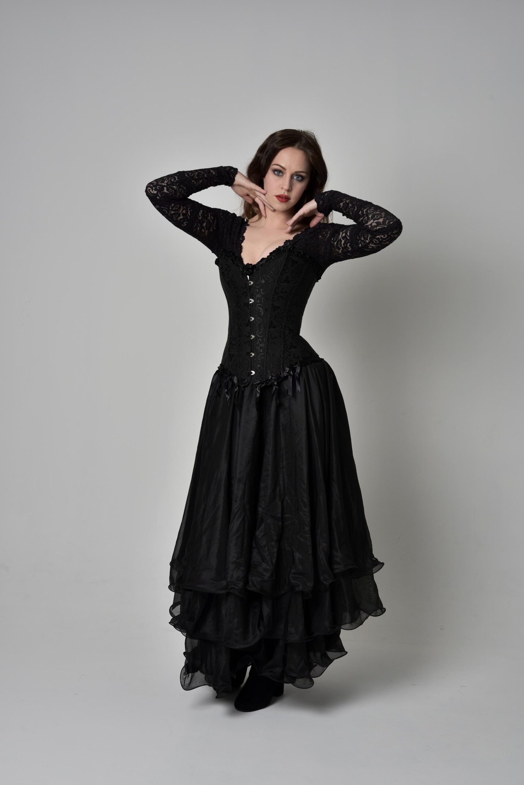 Woman in long black gothic dress and lace sleeves posing against white studio background.