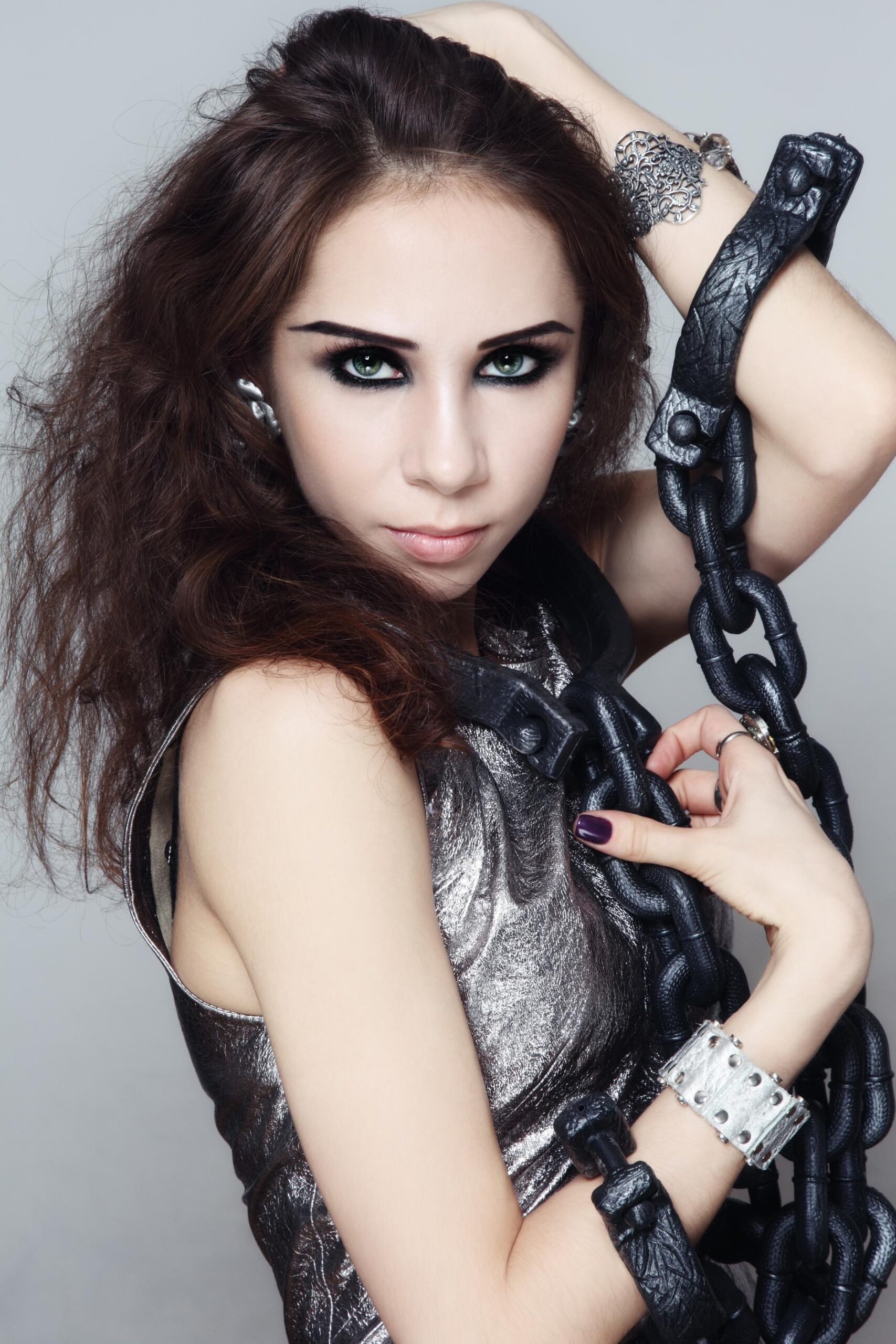 Close-up portrait of woman with smoky eye makeup holding thick black chain against metallic outfit.