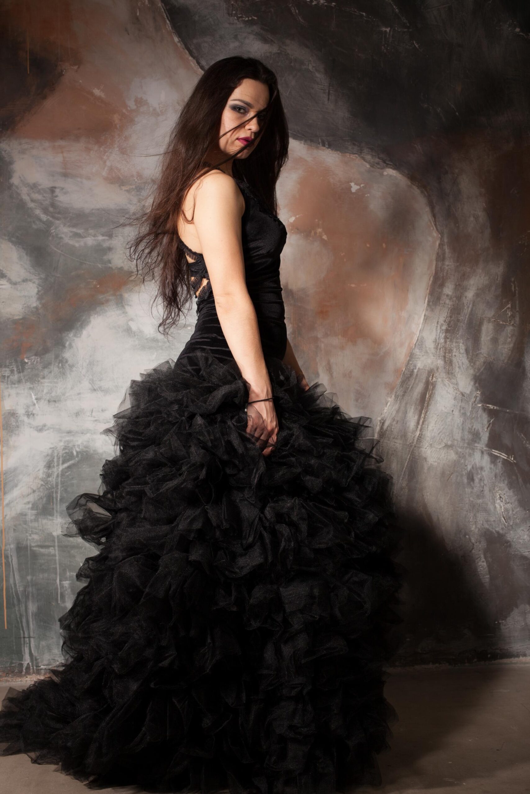 Woman in dramatic black ruffled gown posing against textured studio wall.