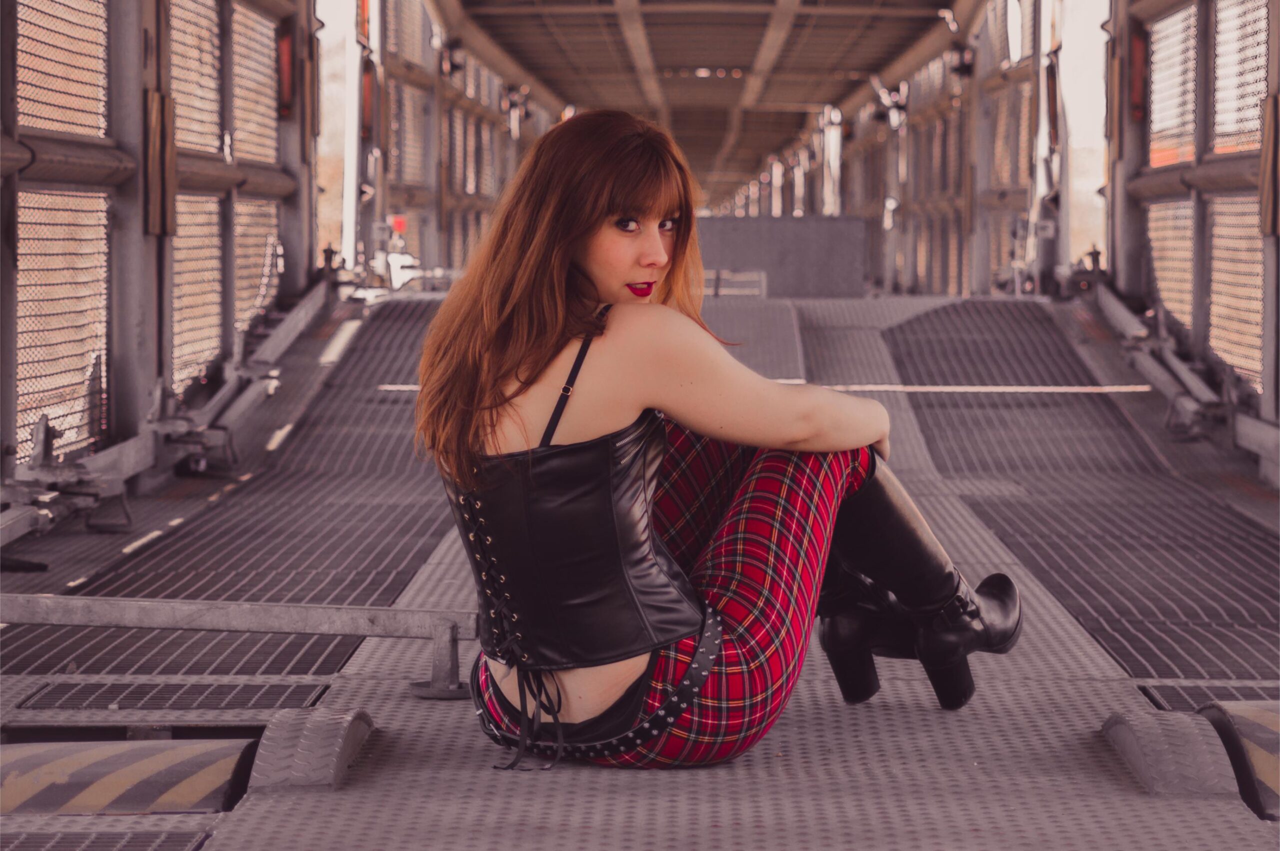 Woman in leather corset and red plaid pants sitting on metal bridge walkway.