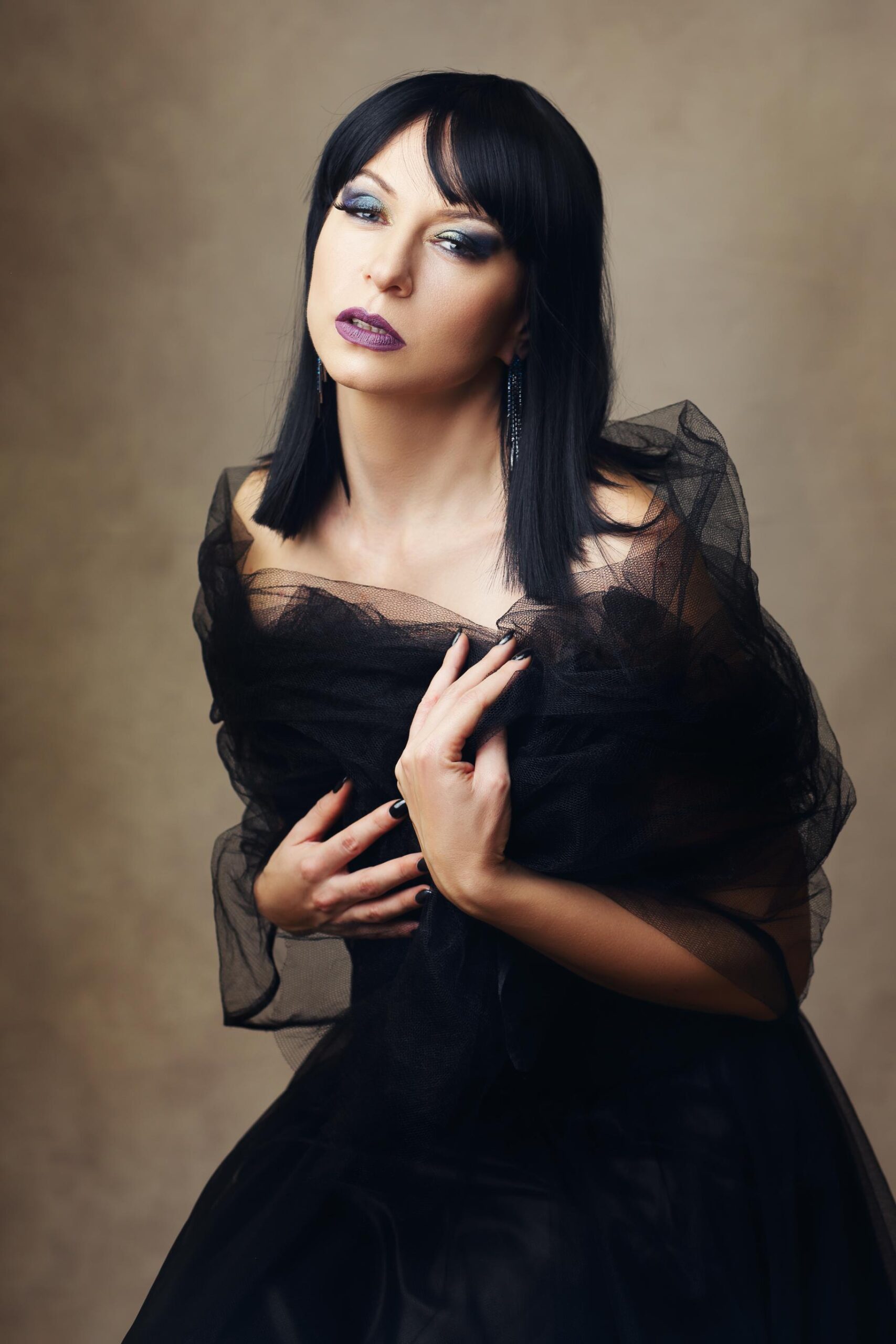 Woman with short black hair and smoky makeup wearing sheer black dress against neutral studio background.