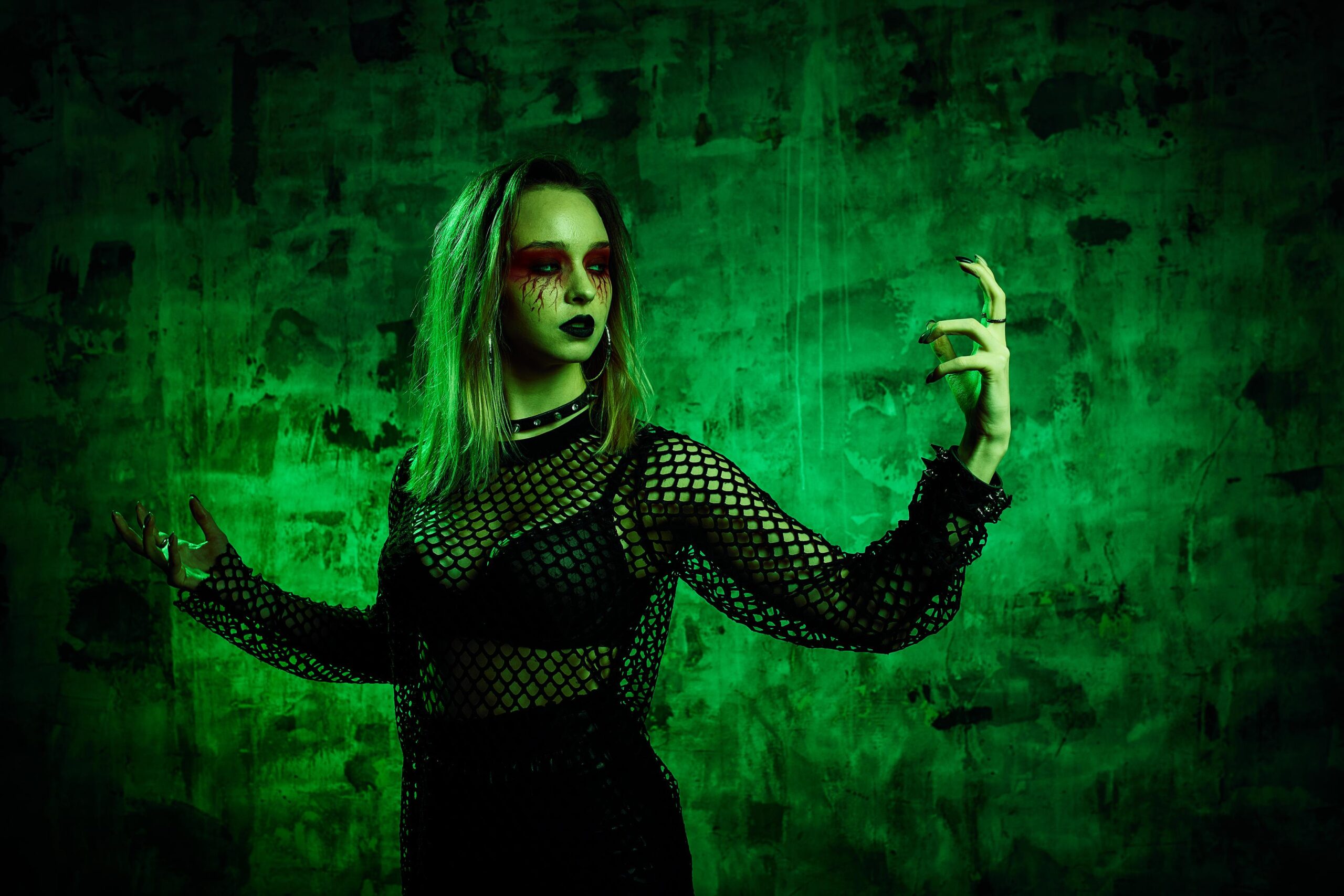 Woman with green lighting and dark makeup wearing fishnet top against grungy wall.