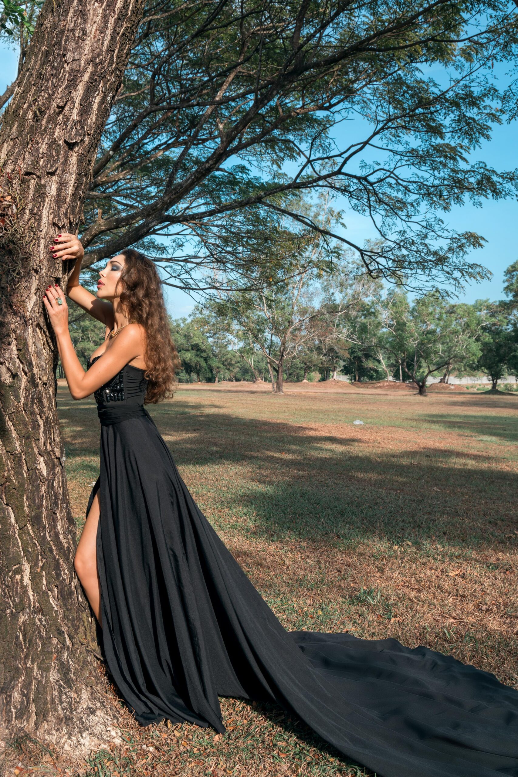 Woman in long black dress leaning against tree in sunny park with flowing train. 