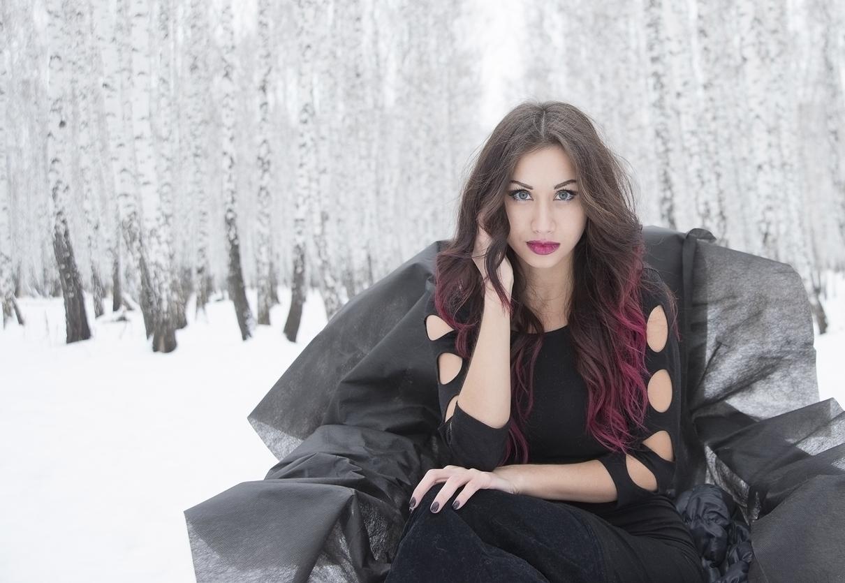Woman with long dark hair sitting in snowy birch forest wearing black dress and intense makeup.