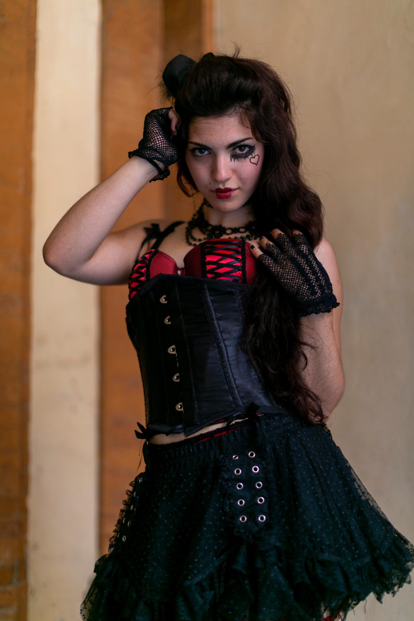 Woman in black and red corset with lace gloves and gothic makeup posing indoors.
