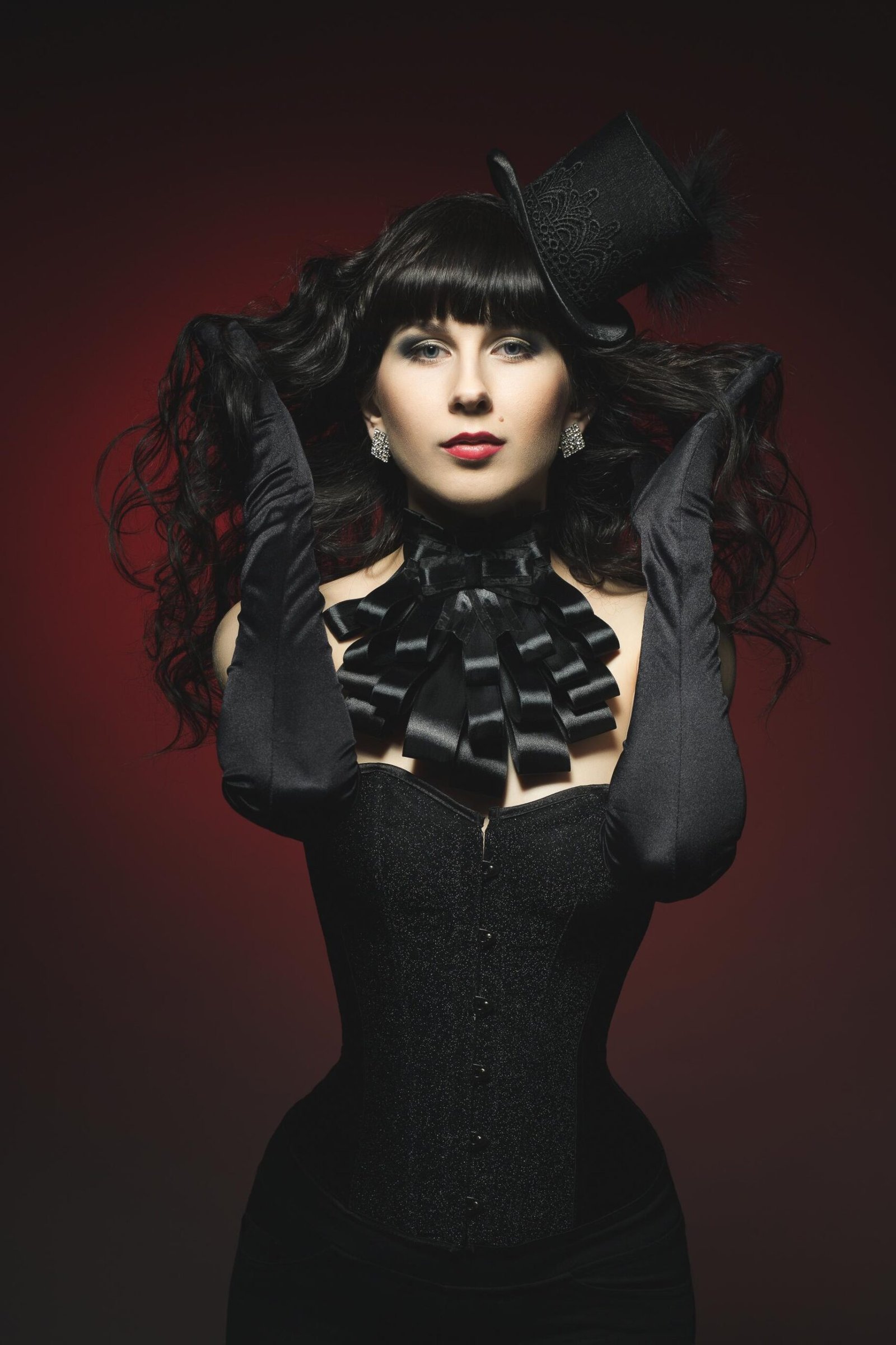 Portrait of woman in black corset and gloves with small top hat on red background.