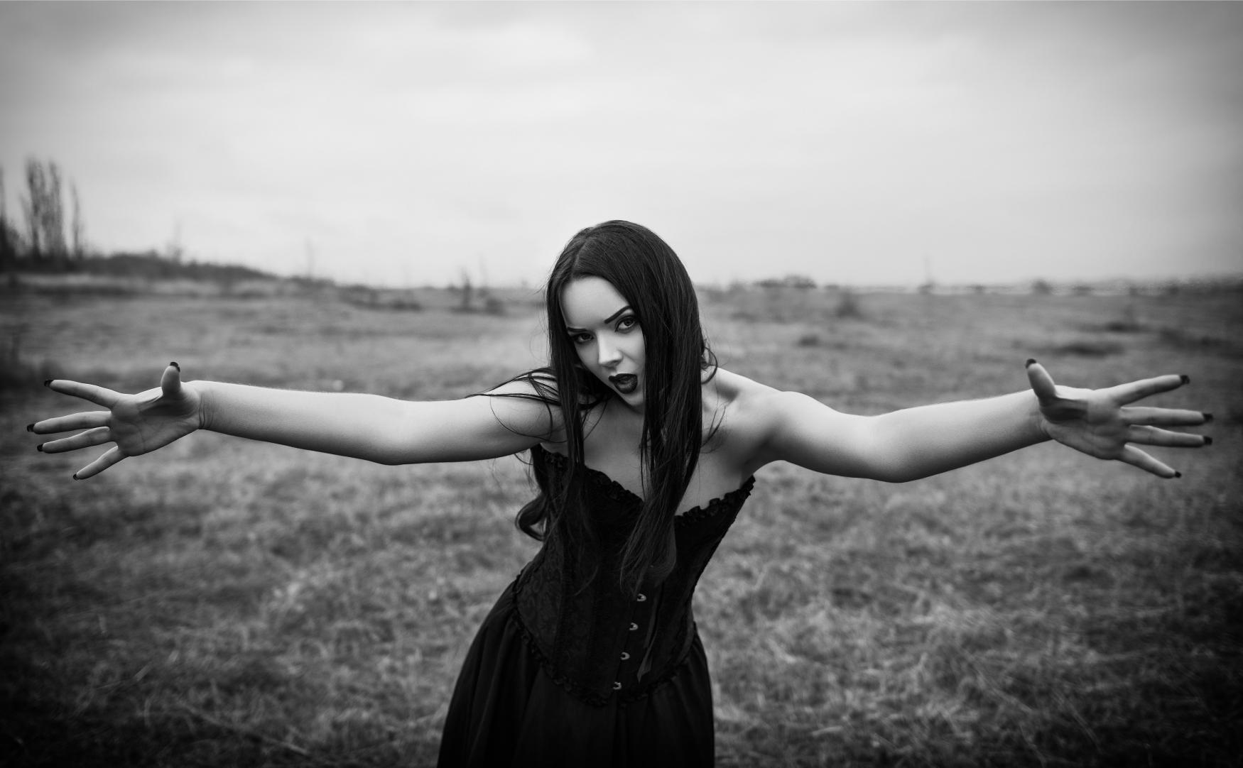 Black and white portrait of gothic woman in corset dress with arms outstretched in open field.