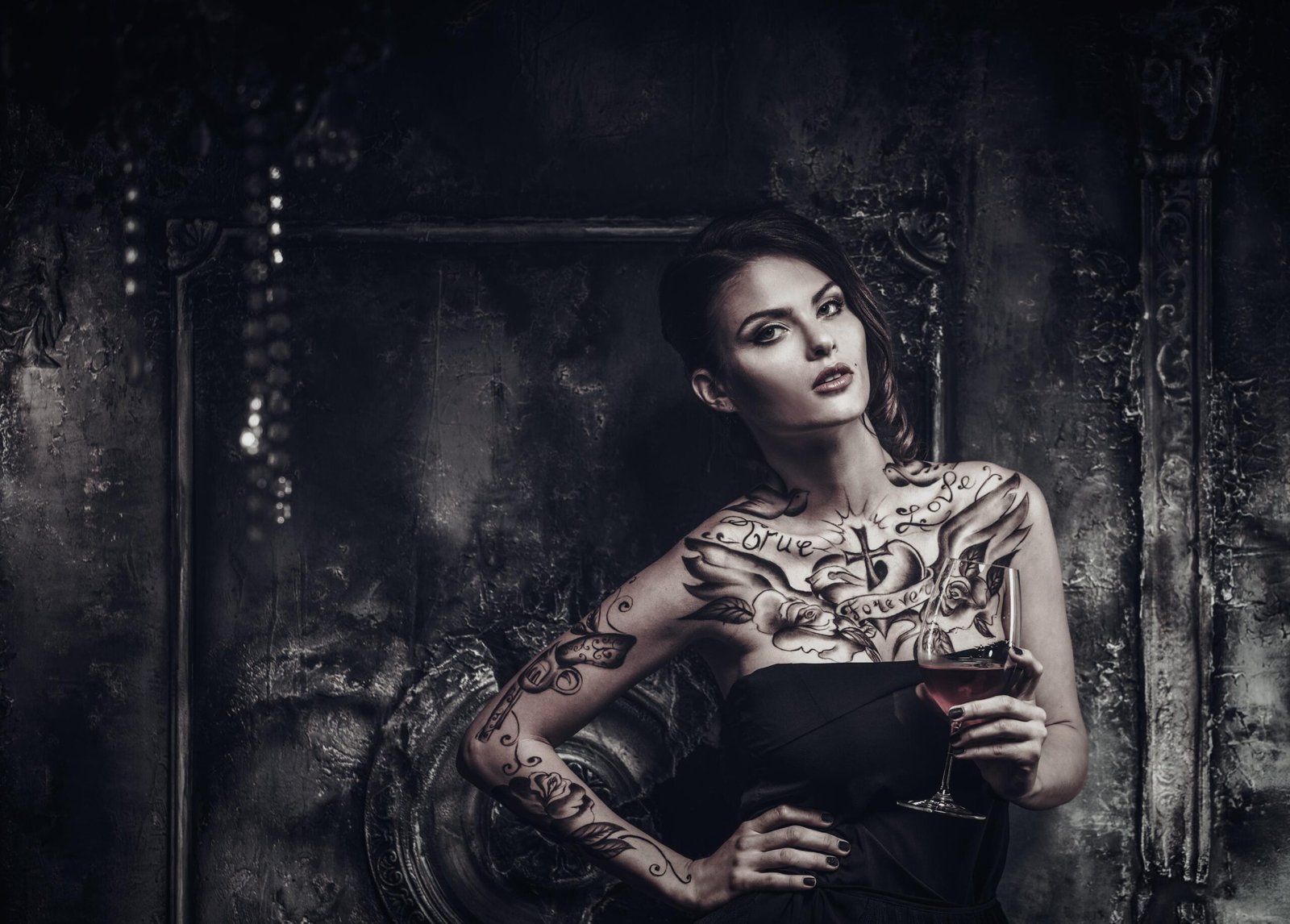 Tattooed woman in strapless black dress holding wine glass in dark vintage room.
