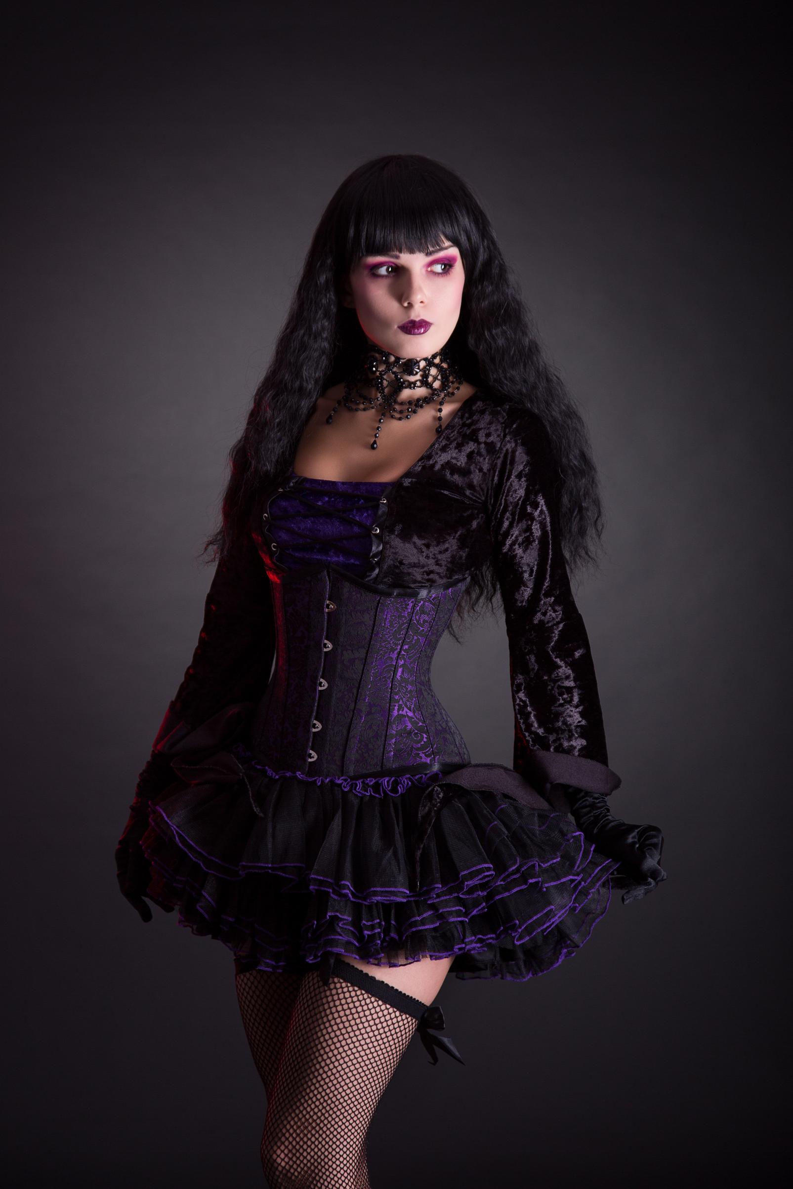 Woman in purple gothic corset dress with fishnet stockings and lace choker posing in studio lighting.