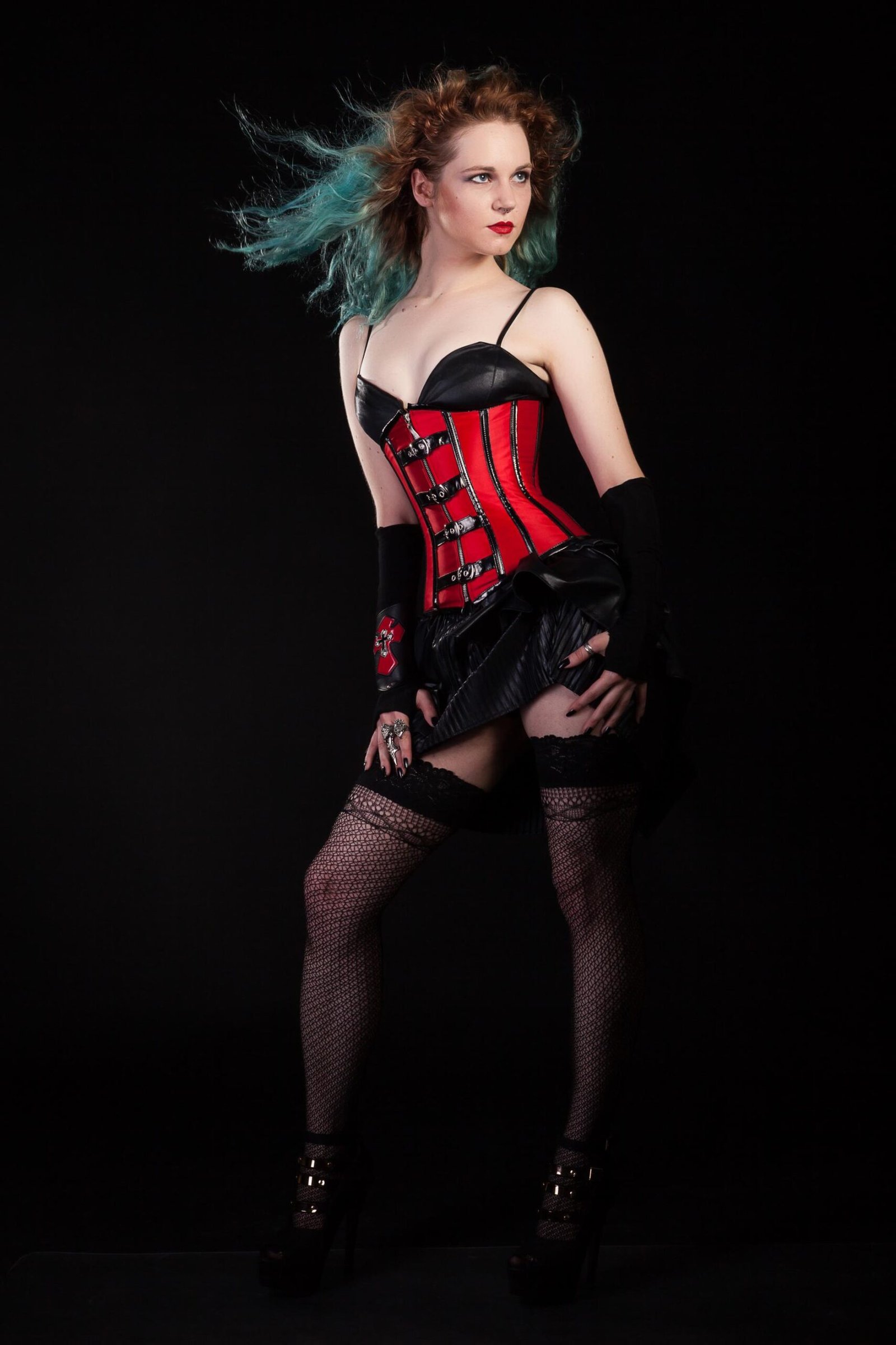 Woman with teal hair in red corset and fishnets posing against black background.