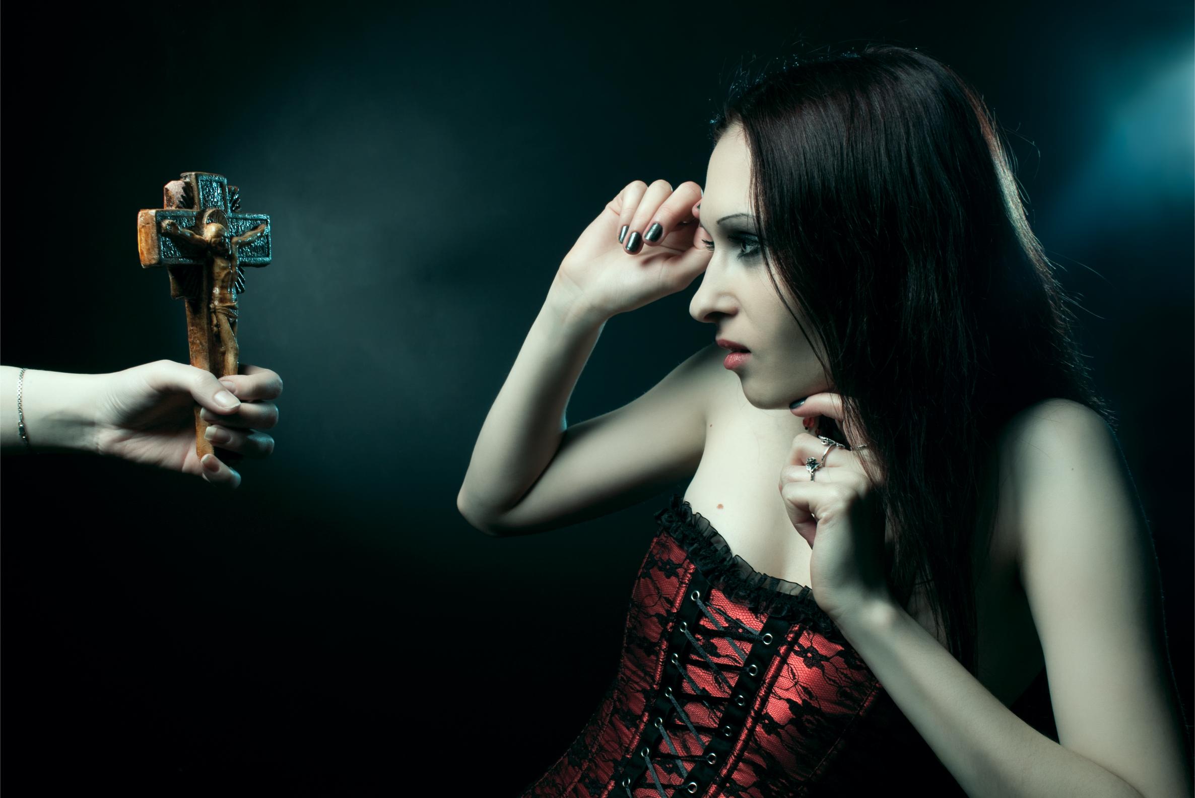 Woman in red corset recoiling from crucifix held toward her in dark dramatic setting.