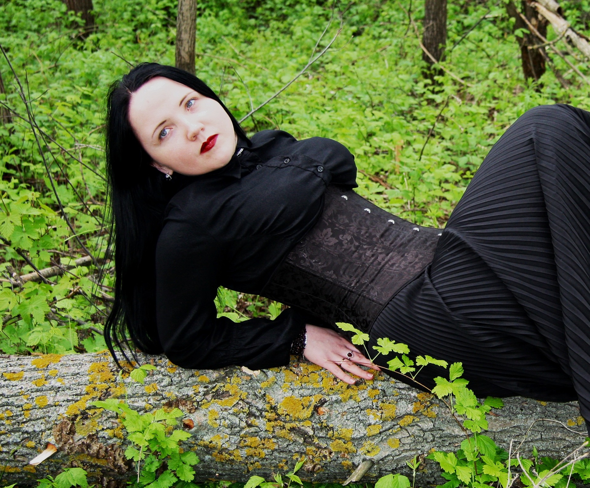 Woman in black gothic outfit reclining on mossy log in green forest.
