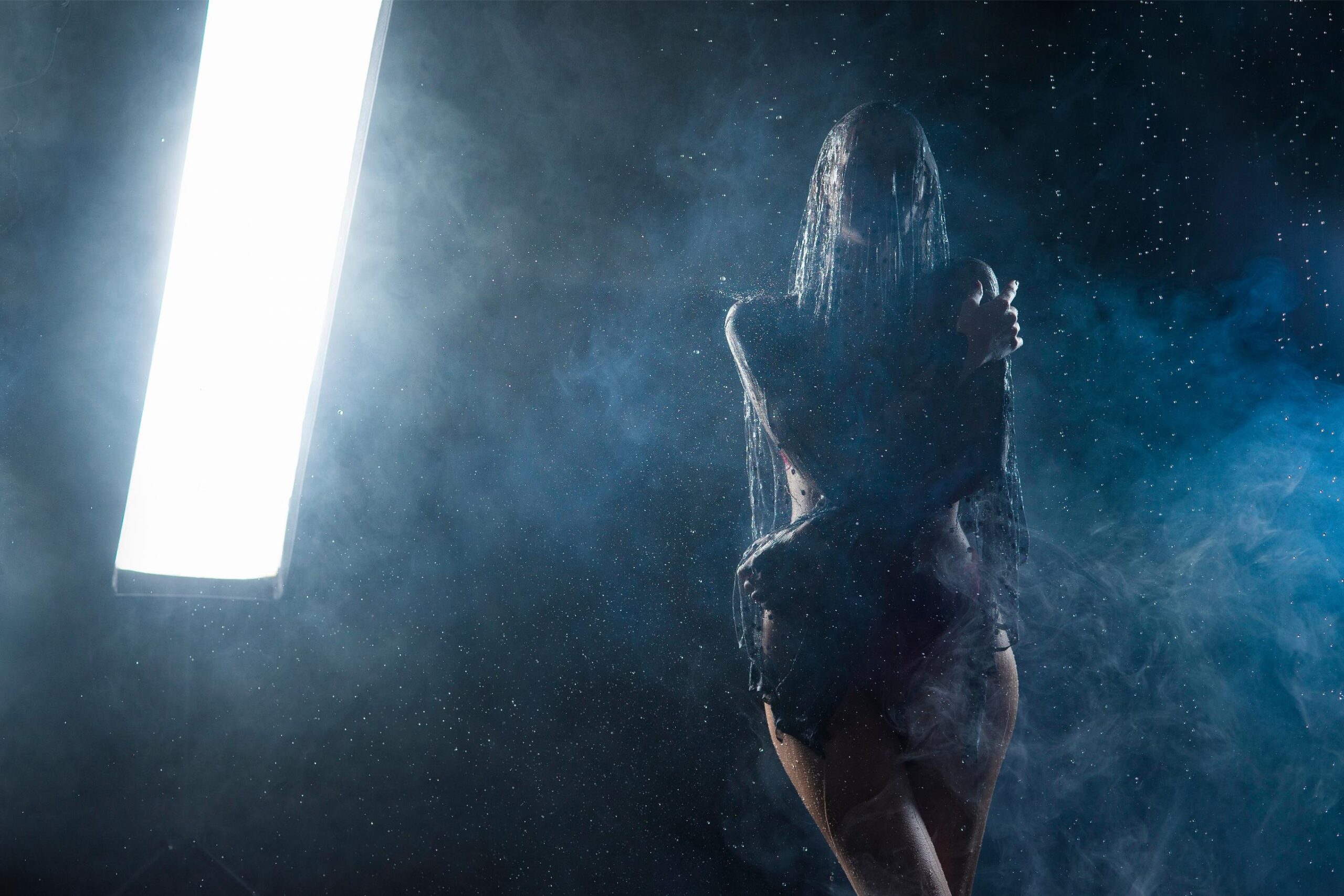 Silhouetted woman standing in mist and light beam with water droplets in dark atmospheric studio scene.