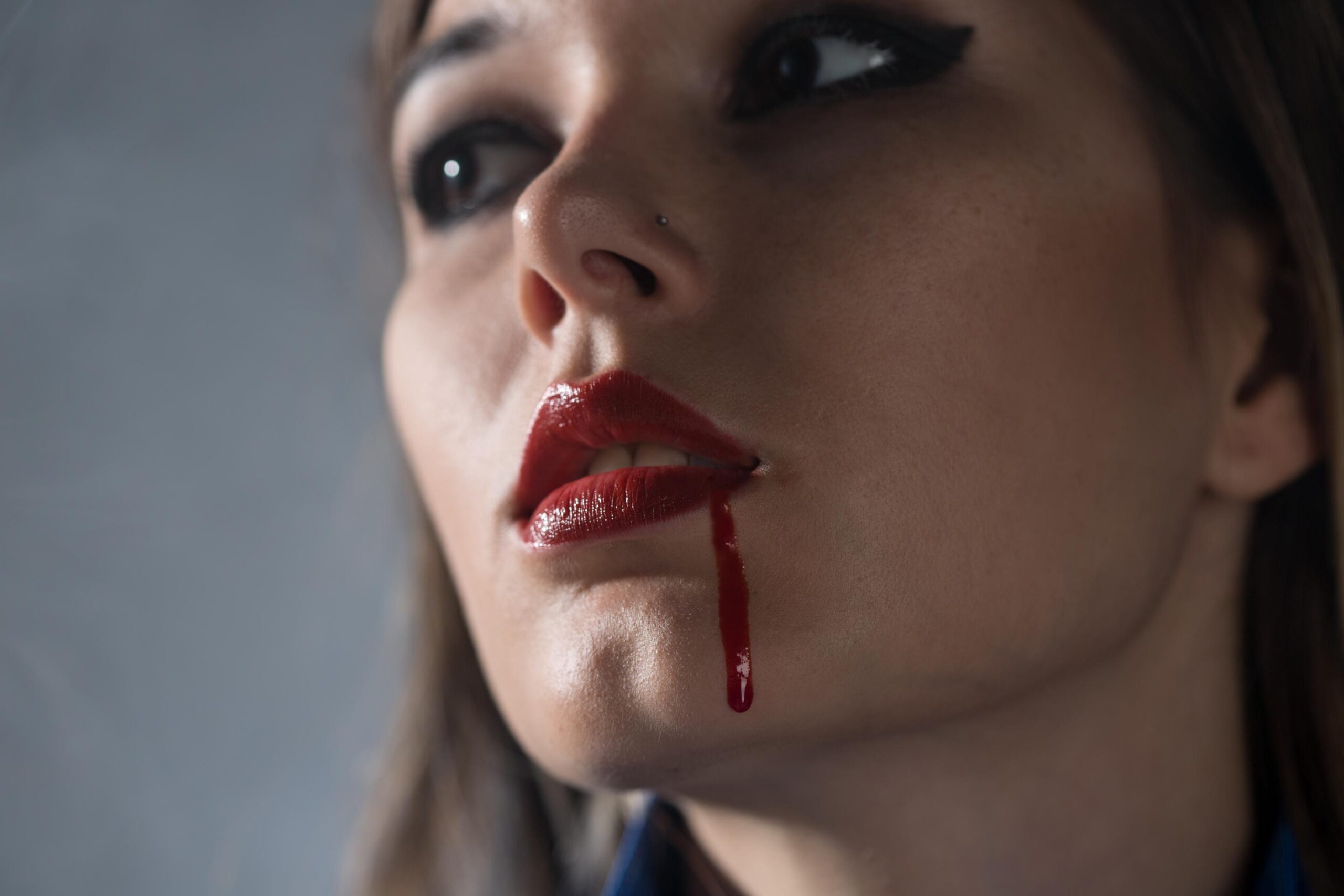 Close-up of woman with red lipstick and blood-like drip from mouth in dark dramatic lighting.