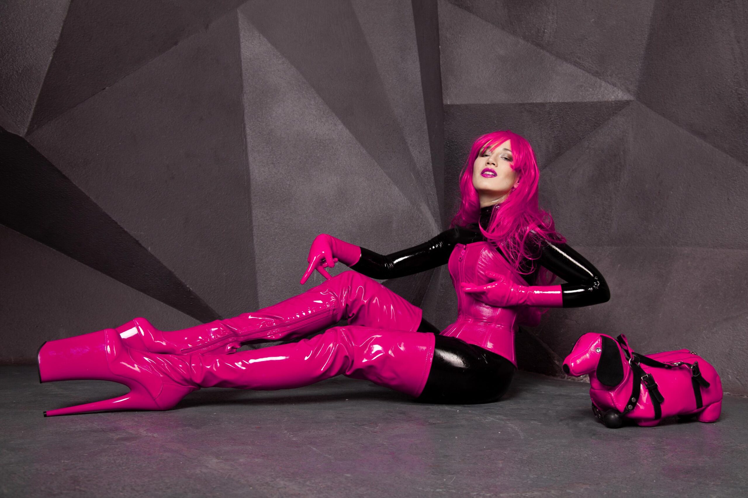 Woman with bright pink hair in glossy pink latex outfit and platform boots posing with matching pink dachshund bag.