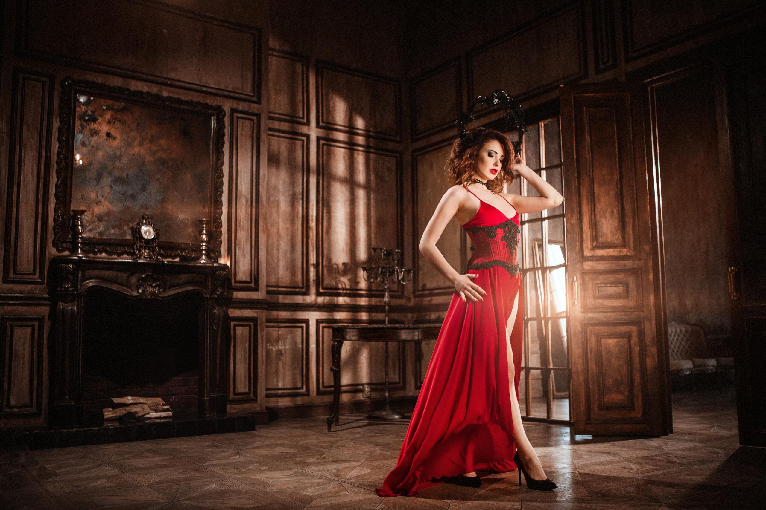 Woman in flowing red dress posing in vintage room with wooden walls and fireplace.