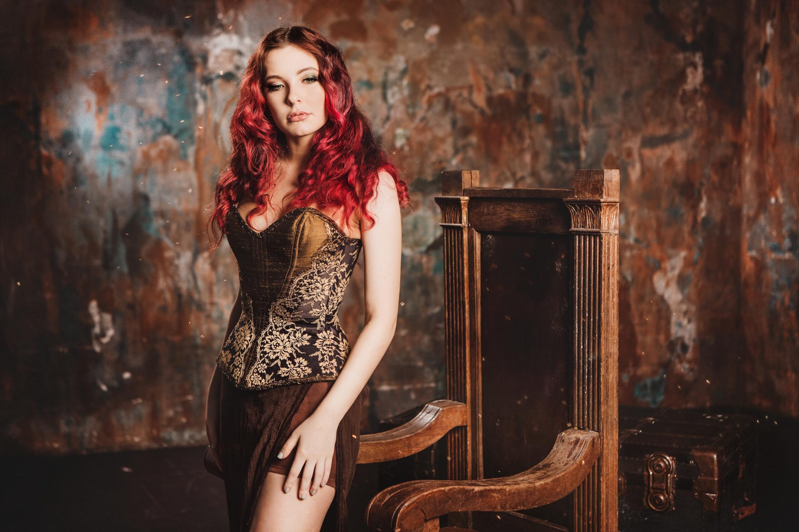 Woman with long red hair wearing ornate brown corset standing beside wooden throne in dramatic studio setting.