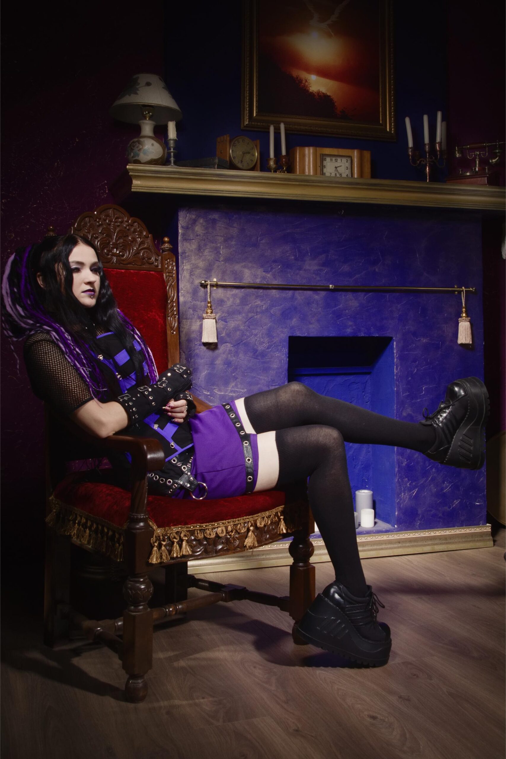 Gothic woman with black hair and purple outfit sitting on ornate chair in vintage room with fireplace.