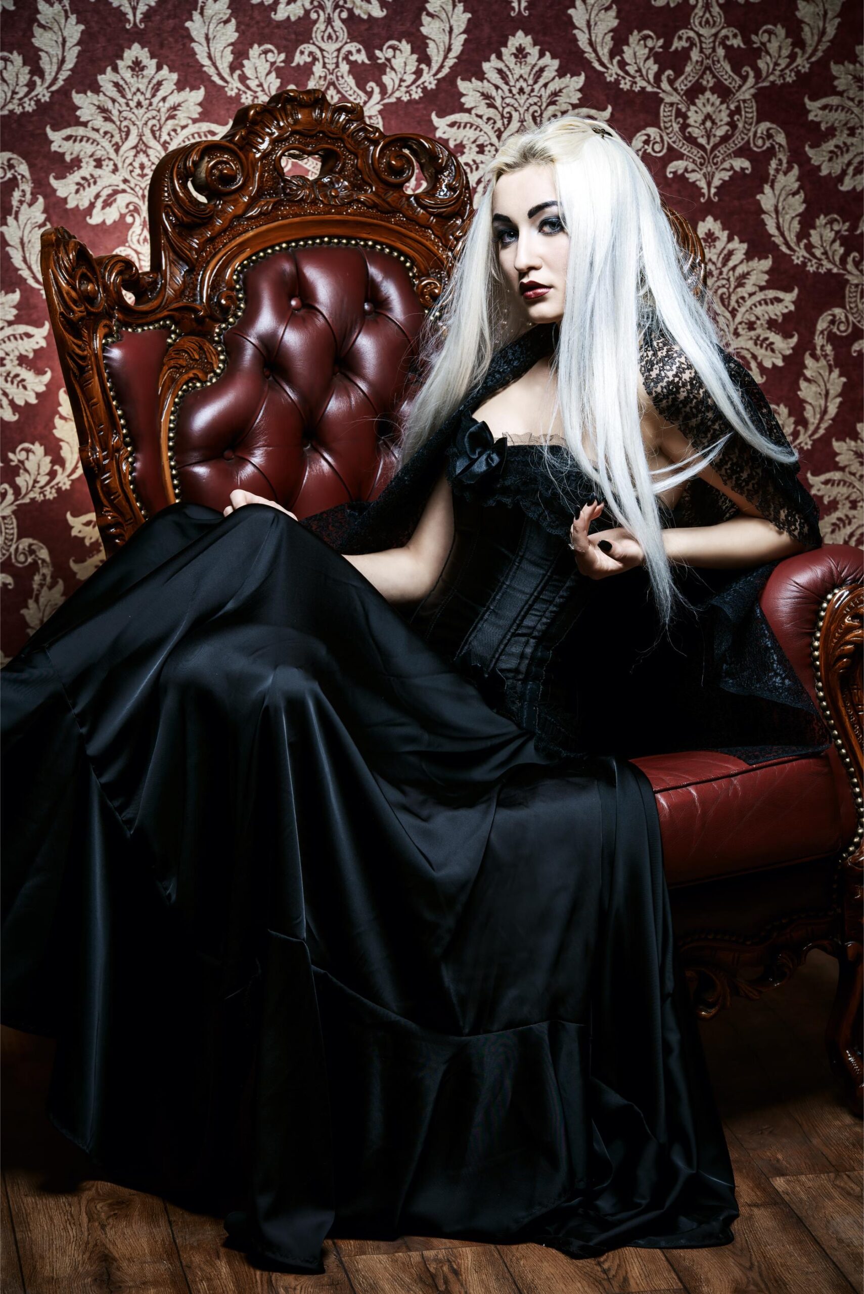Pale woman with long white hair in black gothic dress seated on ornate red leather throne.