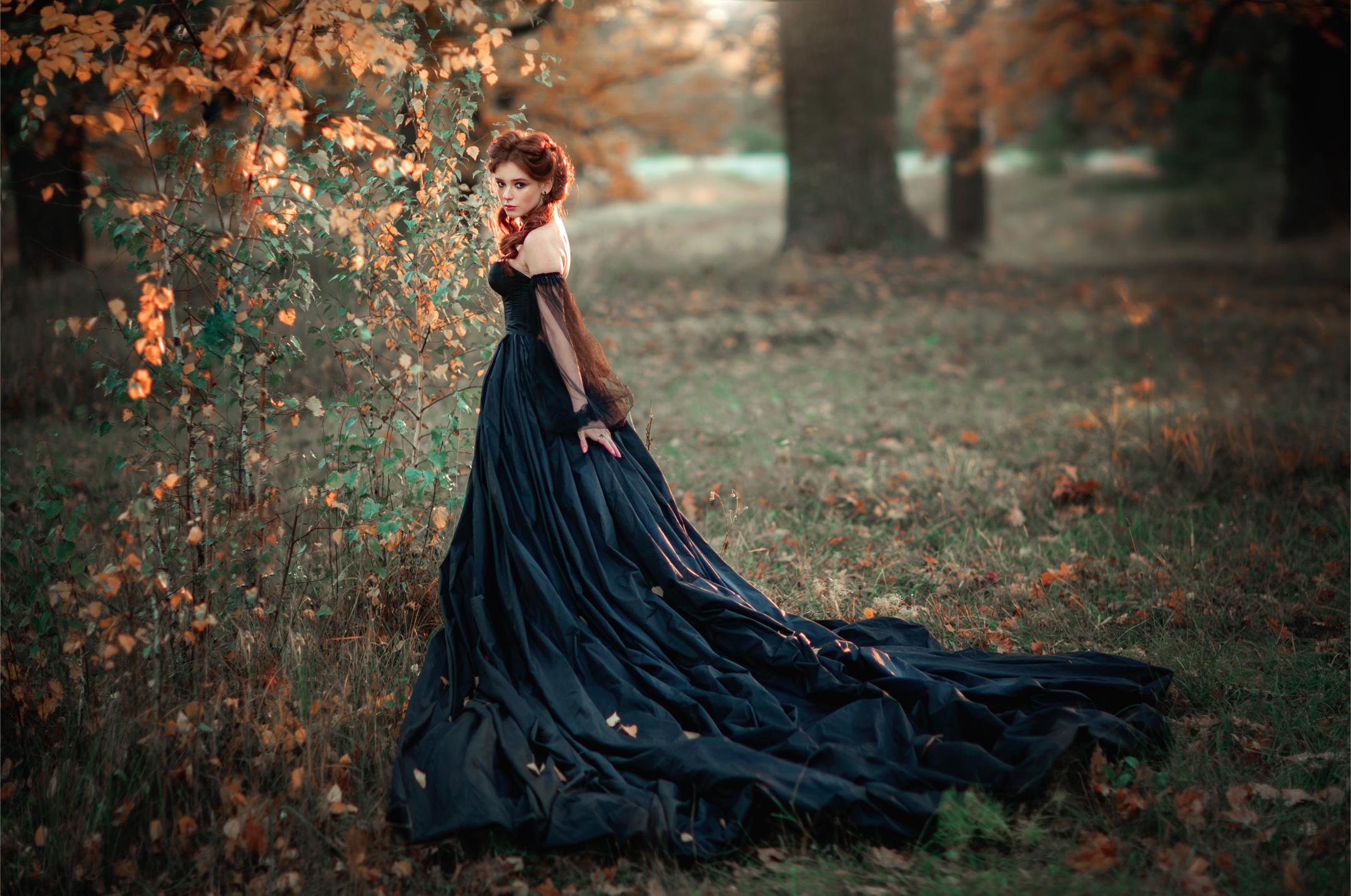 Woman in flowing black gown standing in autumn forest with long dress train among fallen leaves.